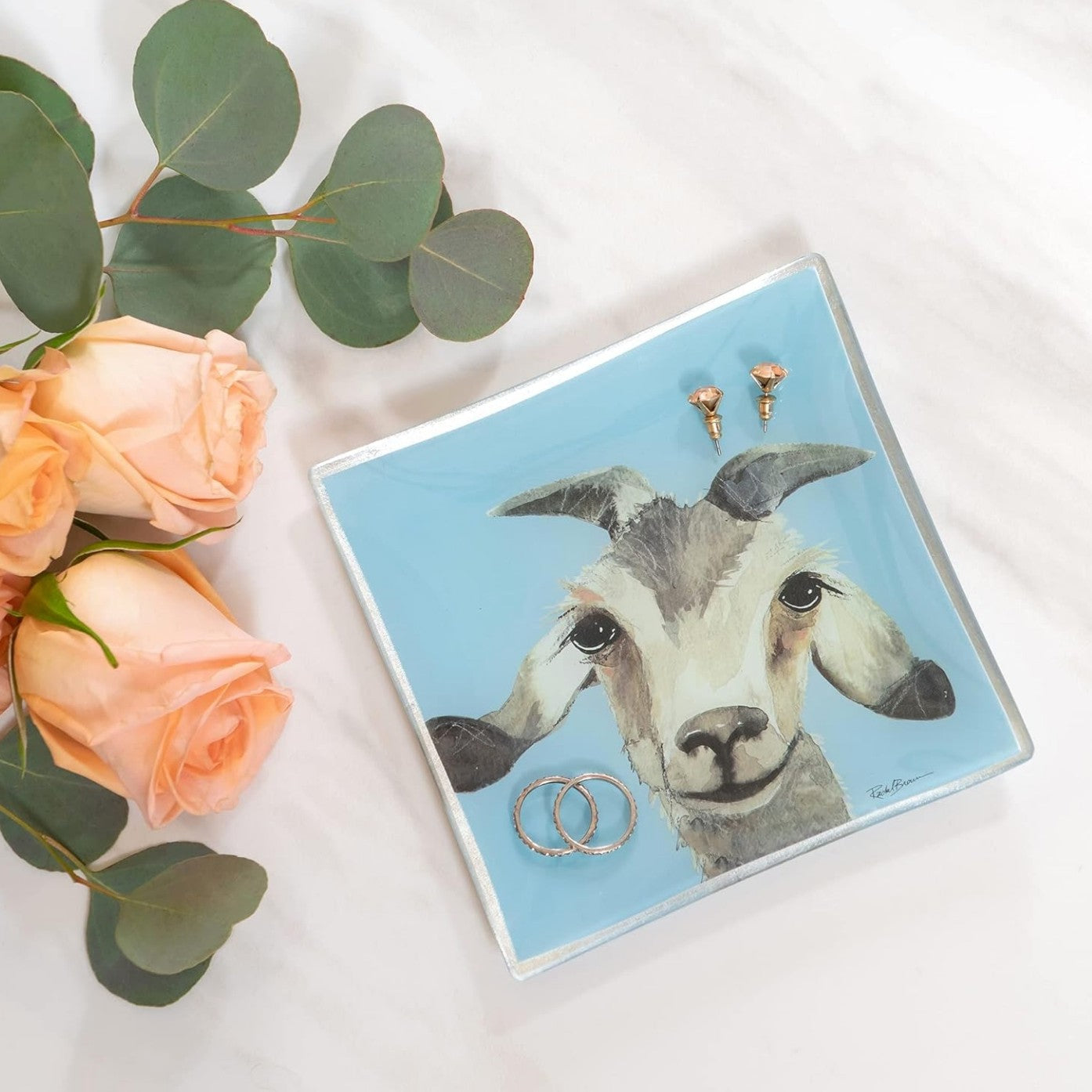 The "Gilbert the Goat" glass trinket dish by Amillia Acres holds two rings and earrings. It sits next to three peach roses and eucalyptus leaves on a white surface.