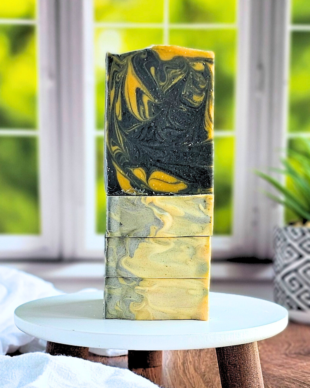 A stack of Amillia Acres' Twilight goat milk soaps, featuring yellow, black, and gray swirls made from natural oils, is displayed on a white pedestal against a softly blurred window with a potted plant in the background.