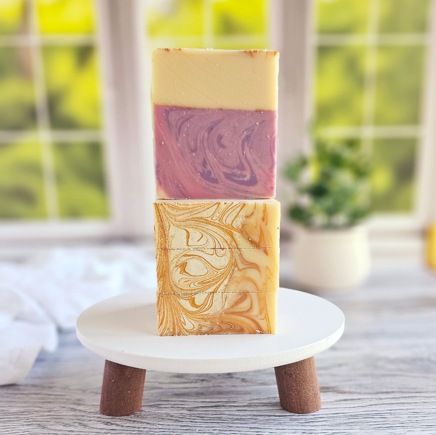 Two Dazzle artisan goat milk soap bars from Amillia Acres rest on a small white pedestal. The top bar features purple and cream marbling, while the bottom has a beige and cream swirl pattern, crafted with natural oils. A blurred window and greenery provide a serene backdrop.
