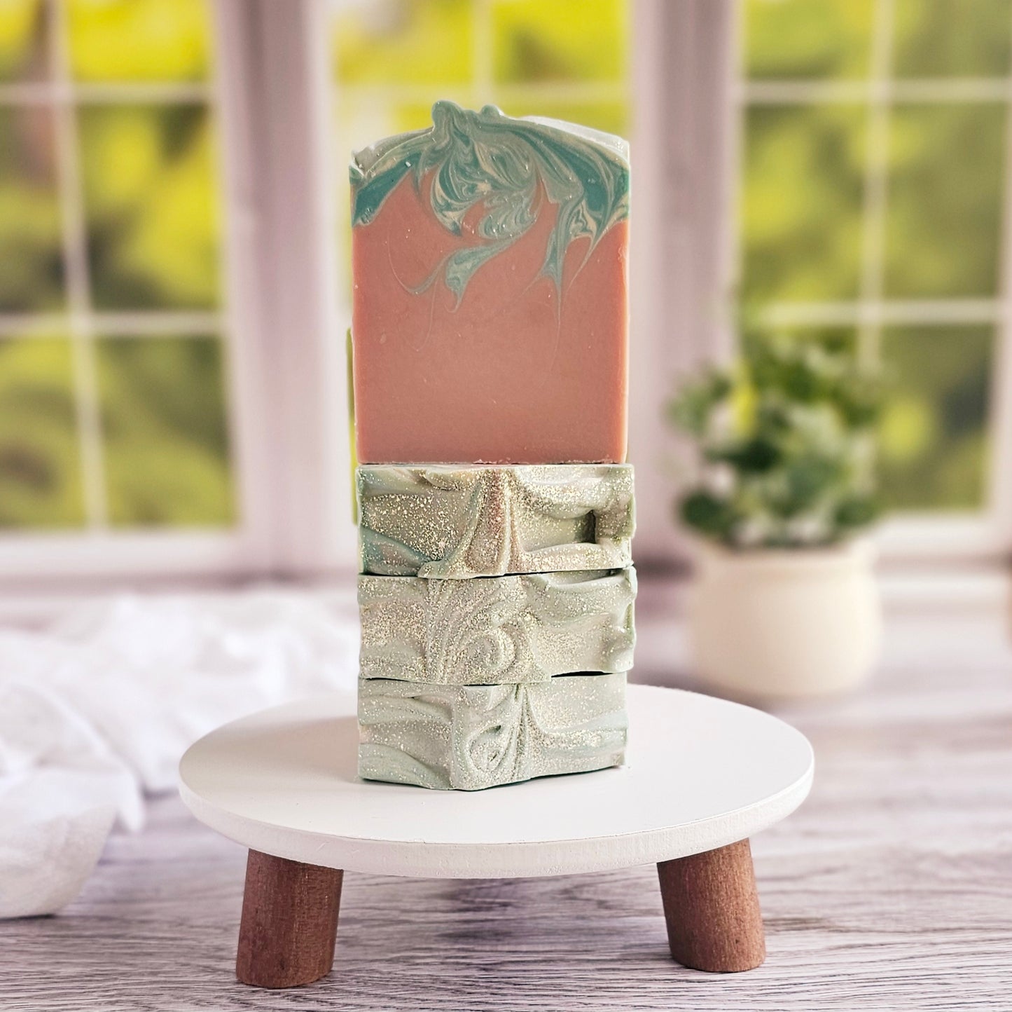 Two Escape artisanal soaps by Amillia Acres, infused with a delightful fragrance, feature swirled patterns and glitter on a white stand with wooden legs. A vibrant window offers a green view, accompanied by a small potted plant—ideal for handmade soap enthusiasts.