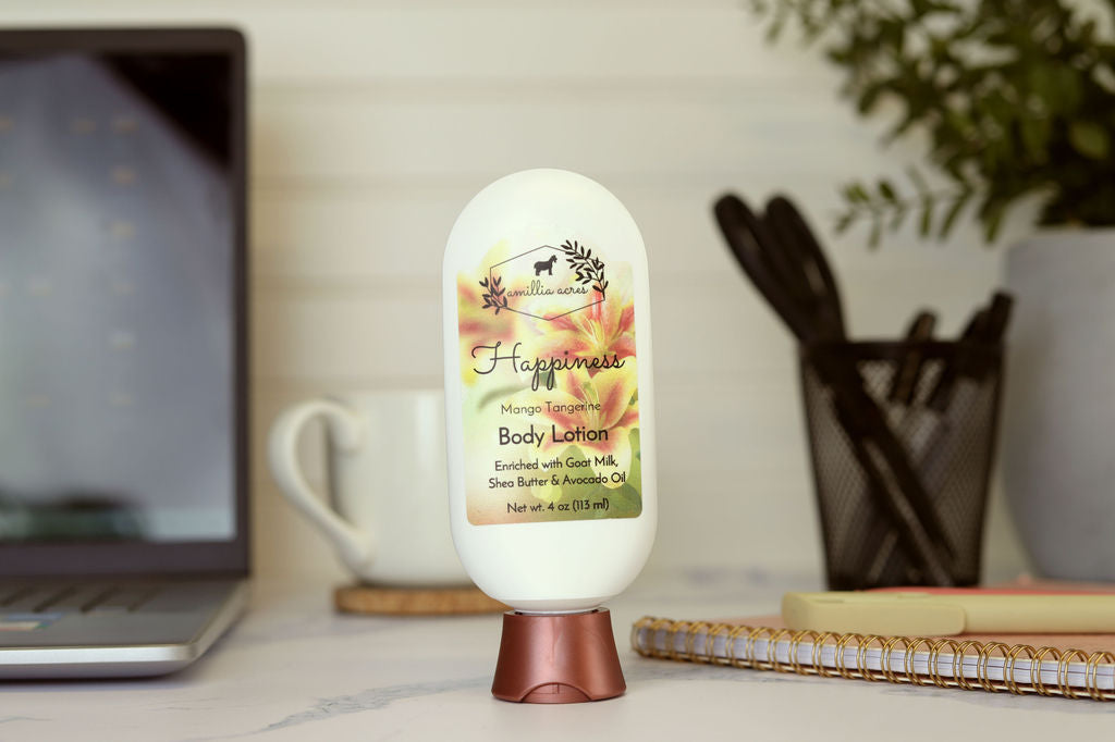 A bottle of Amillia Acres “Happiness” Goat Milk Lotion sits on a desk near a laptop and mug. Its label features nourishing shea butter and avocado oil, offering rich hydration for your skin.