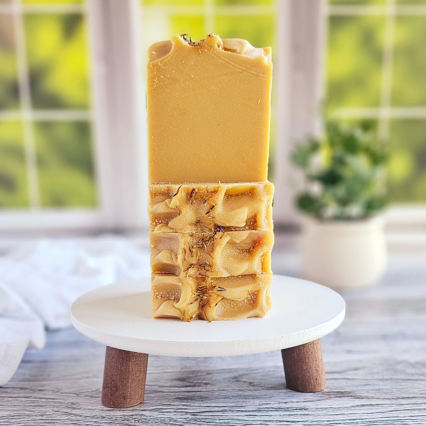 A bar of Amillia Acres' Cheerful goat milk soap, infused with essential oils, rests with a textured rustic charm on a small wooden pedestal. Its top is intricately swirled, set against a blurred backdrop of green foliage and a potted plant near a window.