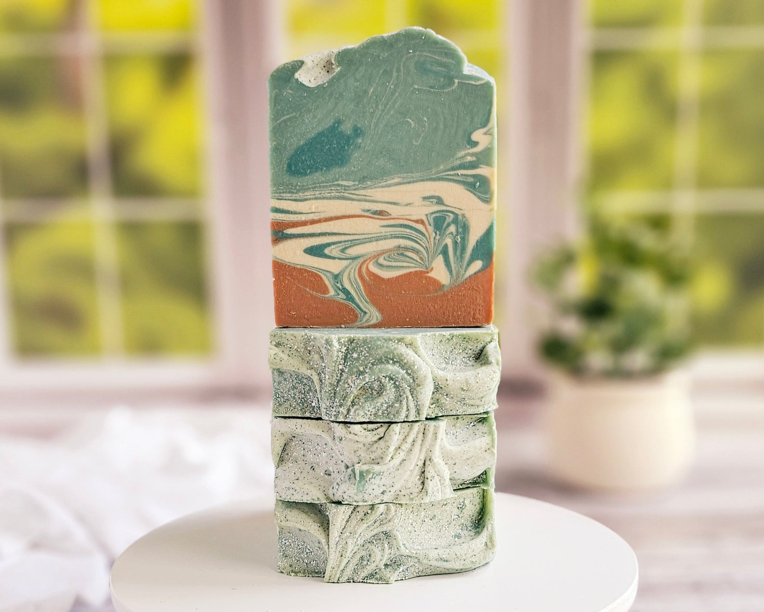 A stack of "Relax" goat milk soaps from Amillia Acres, showcasing swirling green and orange patterns on a white surface. The blurred window and potted plant in the background emphasize the natural oils used in crafting these exquisite bars.