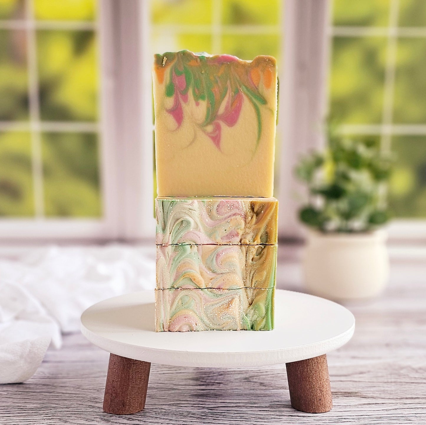 Three bars of Amillia Acres' Happiness Goat Milk Soap, featuring marbled patterns in green, pink, and gold hues, are stacked on a small white stand. Crafted with nourishing natural oils, the scene is set against a blurred window view and a potted plant.