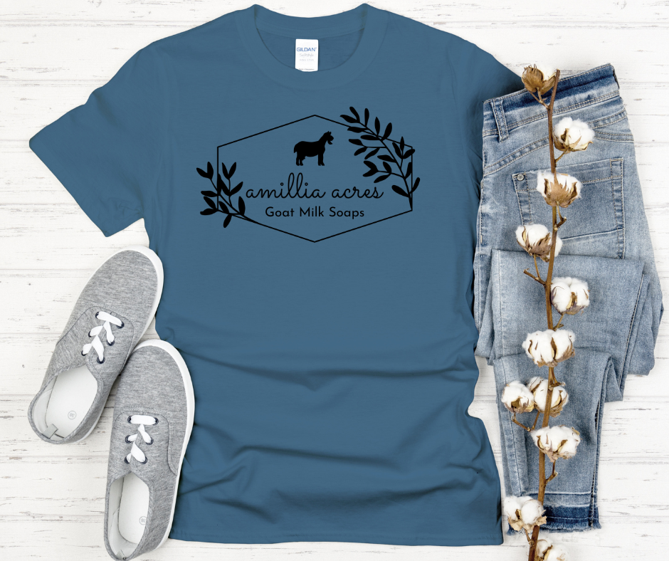 The Amillia Acres ~ Goat Milk Soaps and Lotions short sleeve t-shirt, made from 100% preshrunk cotton, features a charming goat and floral design. It effortlessly complements gray sneakers, faded blue jeans, and a natural touch as it sits gracefully on a white wooden background.