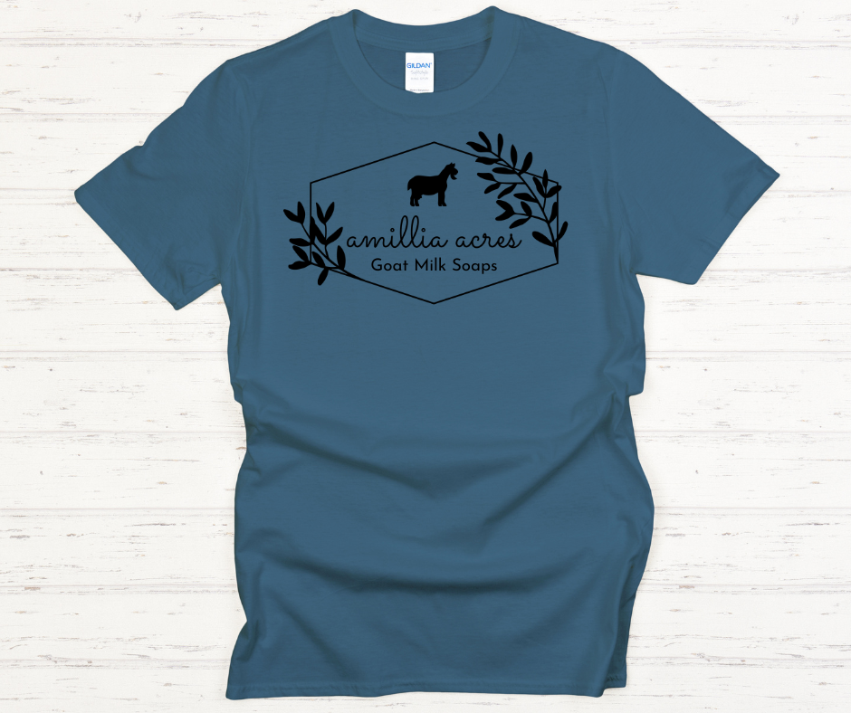 The Amillia Acres ~ Goat Milk Soaps and Lotions T-shirt, crafted from 100% preshrunk cotton, showcases a Gildan Softstyle fit in blue with a black goat silhouette set among leaves. It features the text "Amillia Acres Goat Milk Soaps" within a hexagonal frame and is displayed on a light wooden surface, combining comfort with rustic charm.