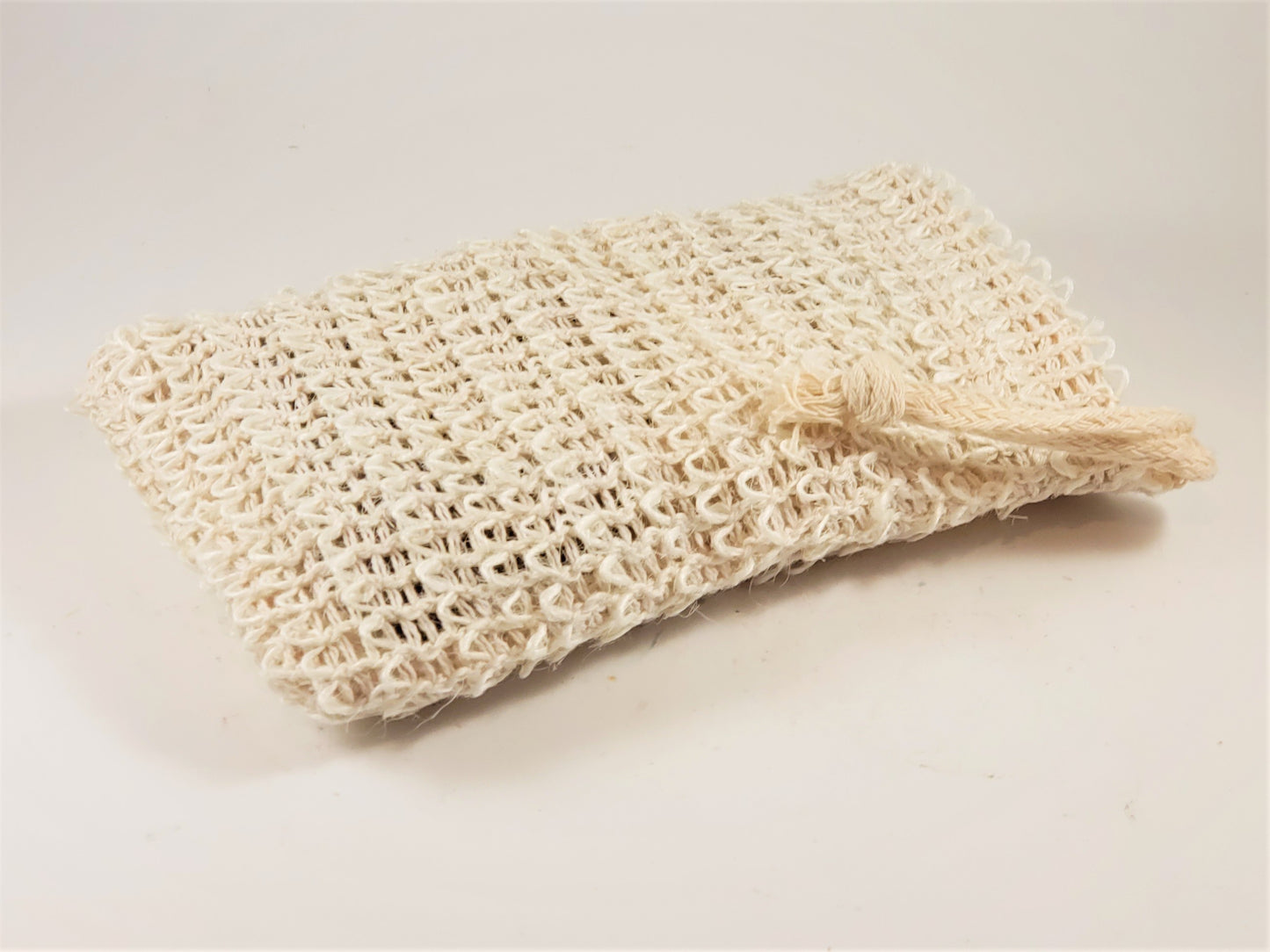 On a white surface rests the Exfoliating Soap Saver Bag by Amillia Acres, a textured beige scrubber crafted from natural sisal with a convenient looped handle. Ideal for gentle exfoliation, it also serves as an exfoliating soap saver.