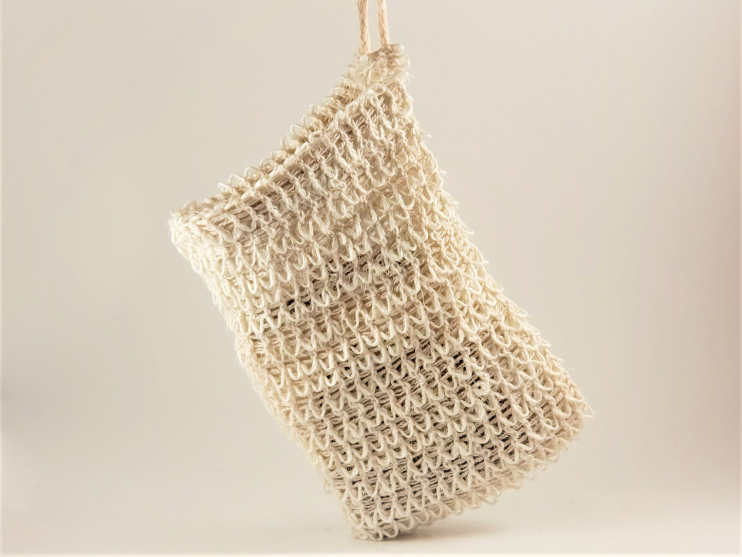The Exfoliating Soap Saver Bag by Amillia Acres, a beige, handwoven mesh-style accessory, is displayed against a plain, light-colored background. Crafted from natural sisal, its texture boasts intricate loops and knots for an airy and natural appearance. Perfect for gently exfoliating your skin during daily routines.