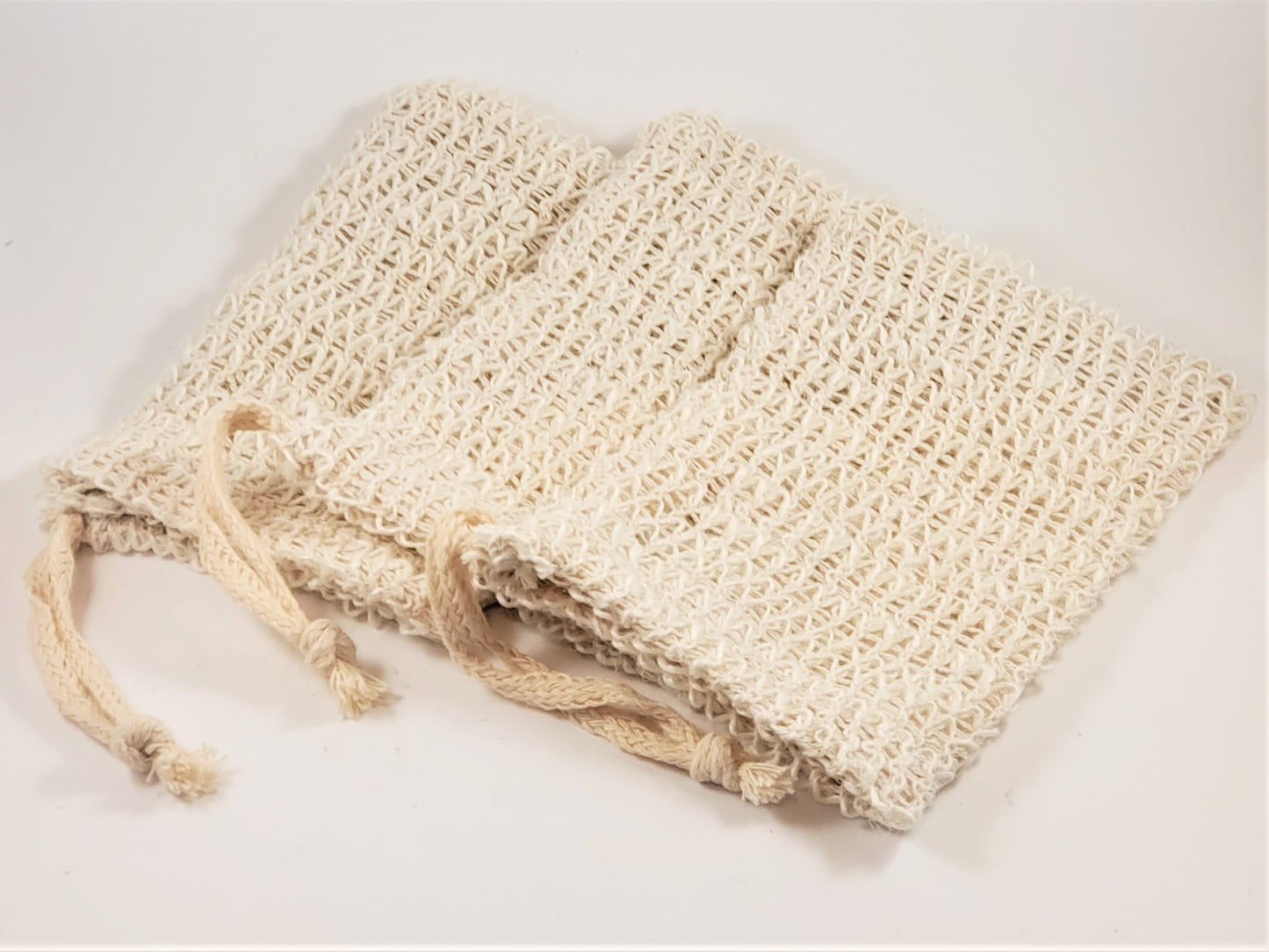 On a light surface lie three Exfoliating Soap Saver Bags by Amillia Acres, crafted from natural sisal in beige. Each bag features a drawstring cord for closure and is designed for gentle exfoliation.