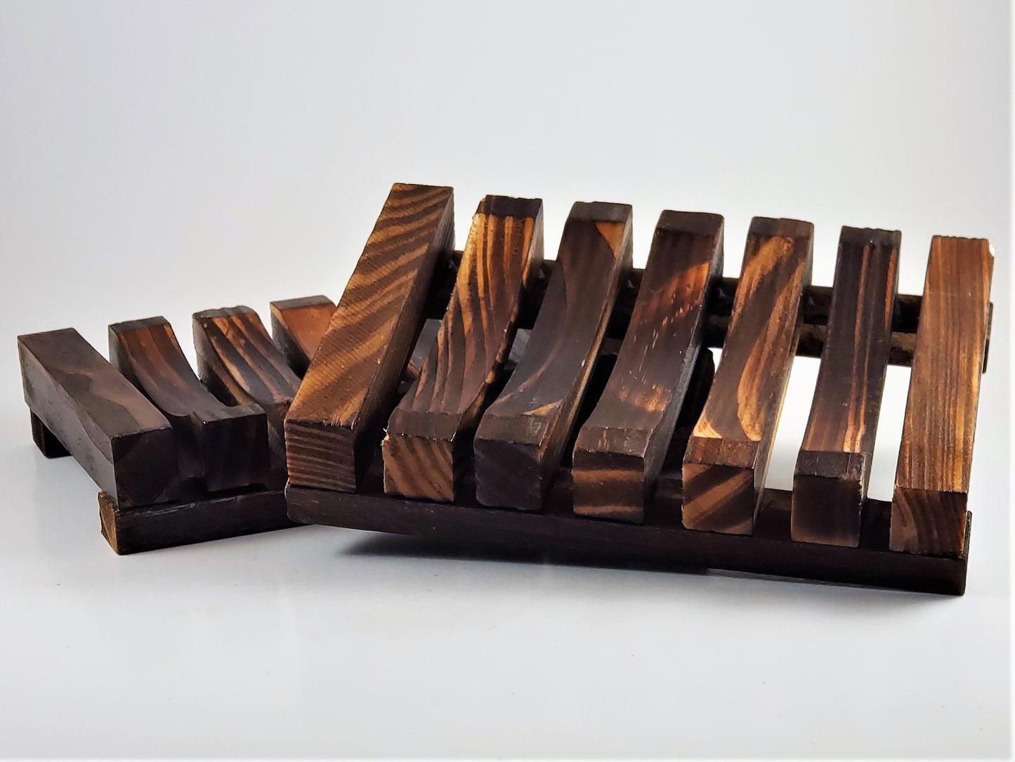 On a white surface, two Amillia Acres Wooden Soap Lifts with slatted designs display their rich brown color and visible wood grain. These rustic soap savers naturally elevate the soap, promoting efficient drying to extend its lifespan.