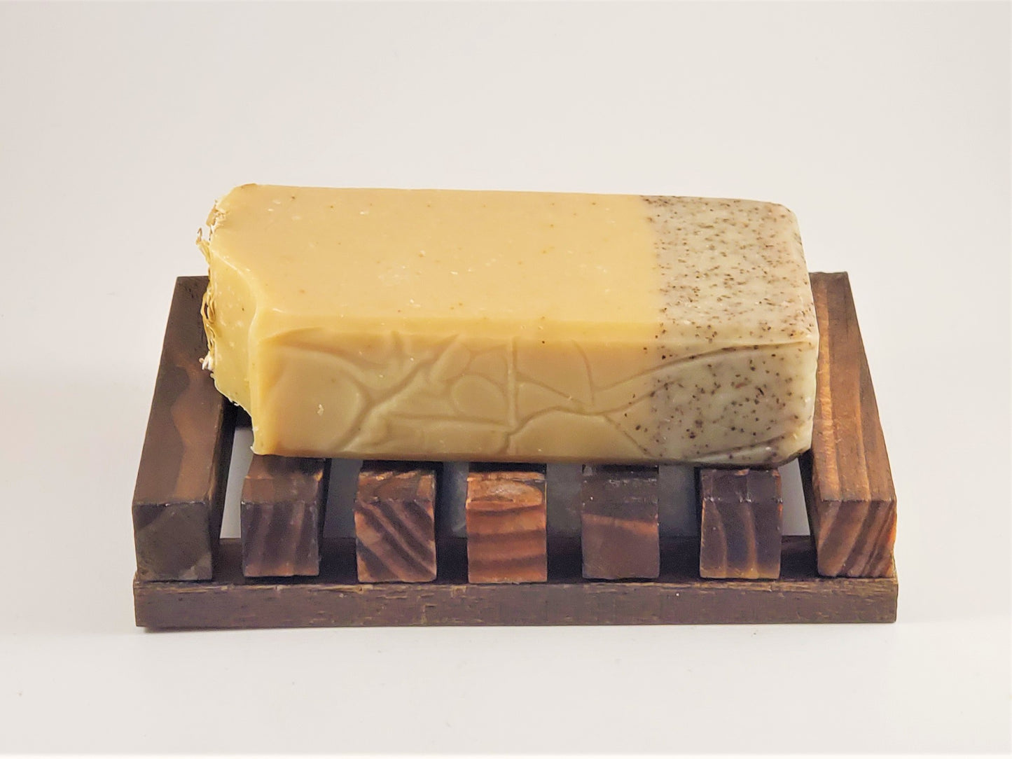 A rectangular bar of soap with a textured, two-toned surface rests on the Amillia Acres Wooden Soap Lift featuring natural wood slats. The soap showcases a section of speckled dark particles for exfoliation against its plain background, elegantly displayed on the wooden lift.