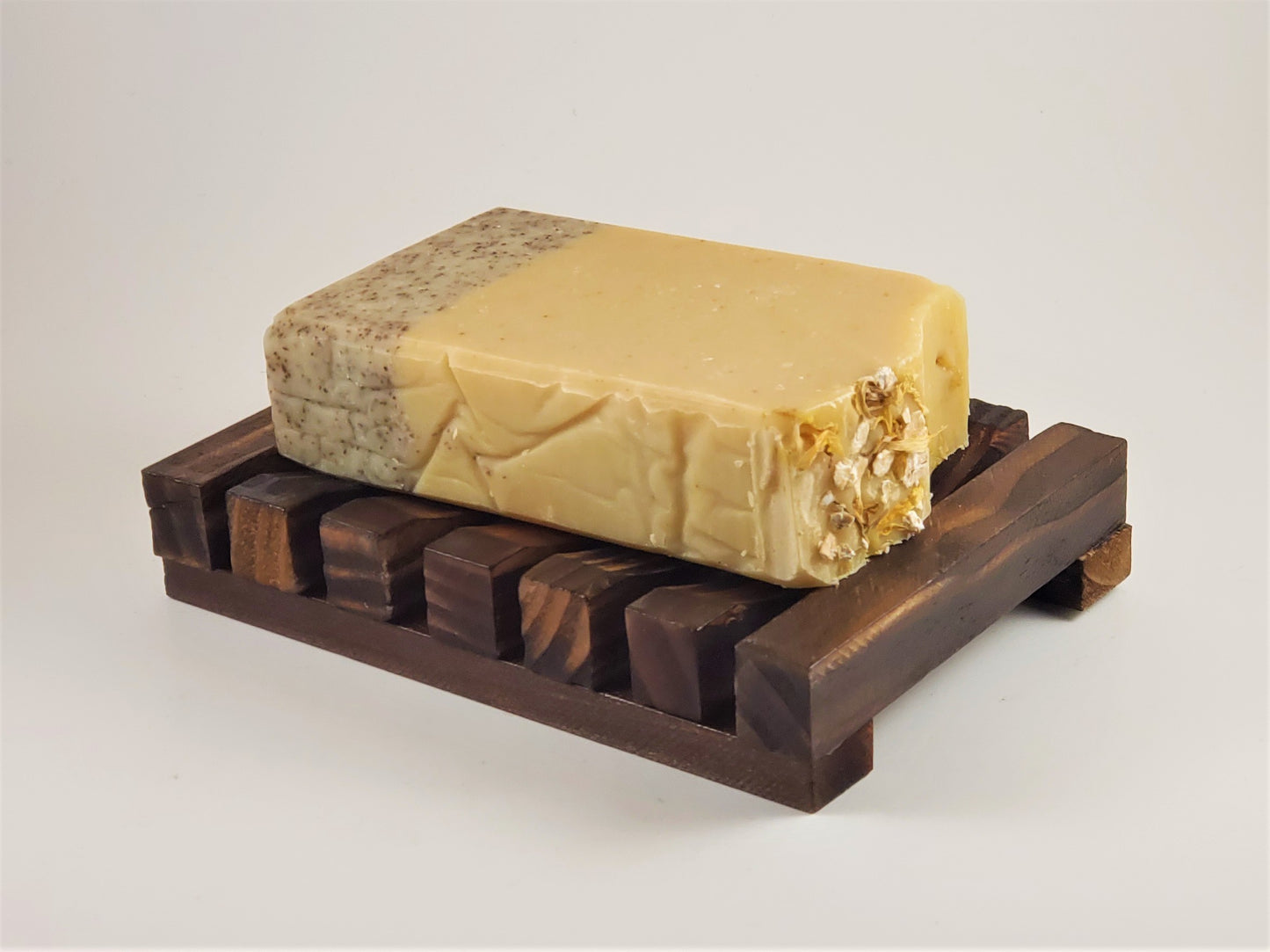 A bar of handmade soap with a textured, two-tone beige design sits elegantly on an Amillia Acres Wooden Soap Lift. Embedded oats on one side provide a rustic charm, all set against a plain white background.