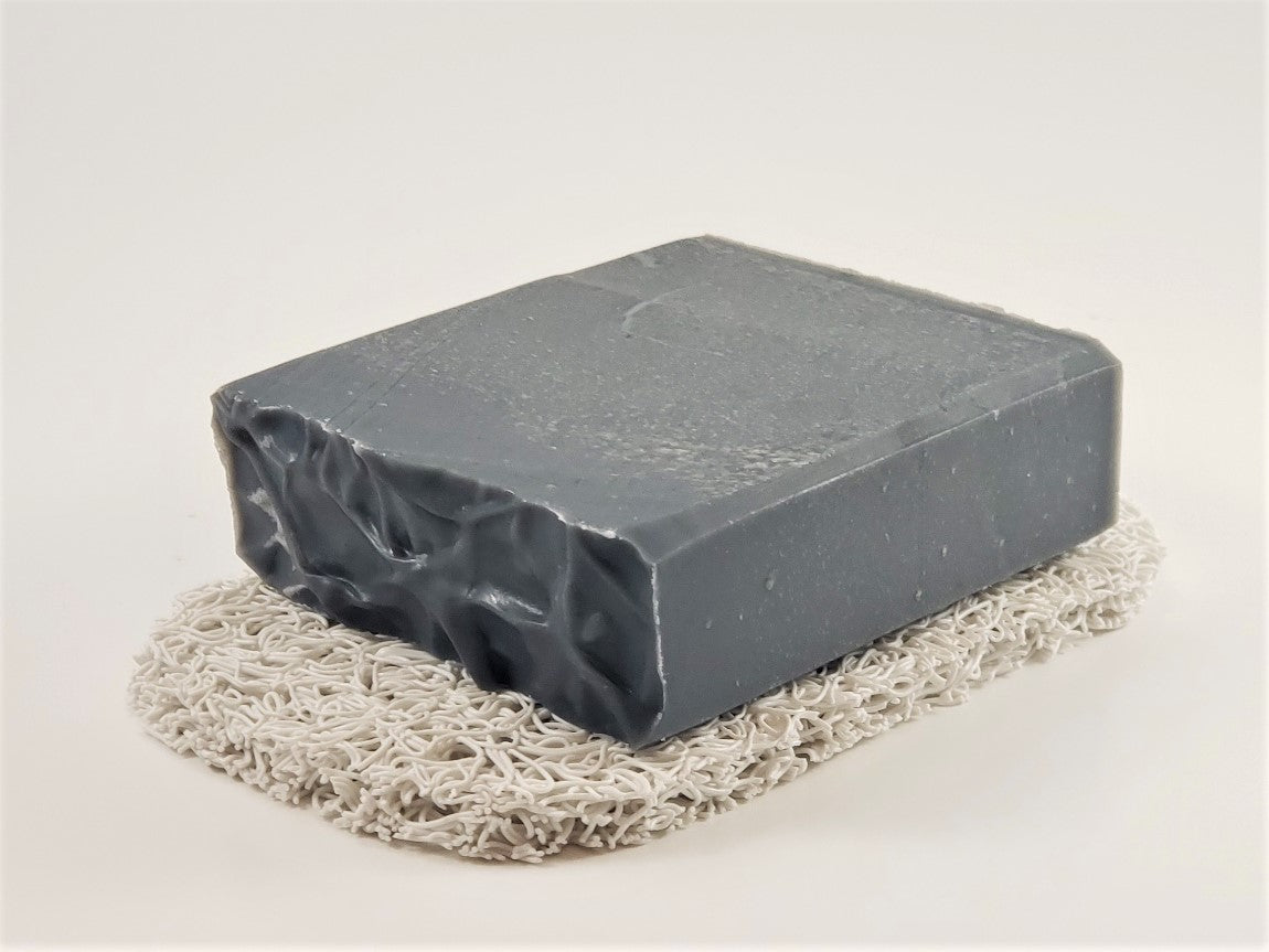 A dark gray, rectangular bar of soap with a rough, textured surface on one side is placed on the Amillia Acres "Soap Saver," a light, woven mat, set against a plain white background.