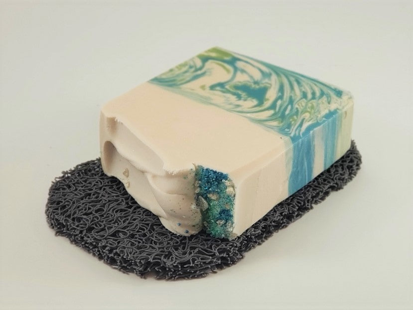A decorative soap bar featuring blue and green swirls with a textured gray base is showcased on Amillia Acres' Soap Saver, a dark and curly mat designed to extend the life of your soap. The visible edge reveals the bar's layered design and smooth finish.
