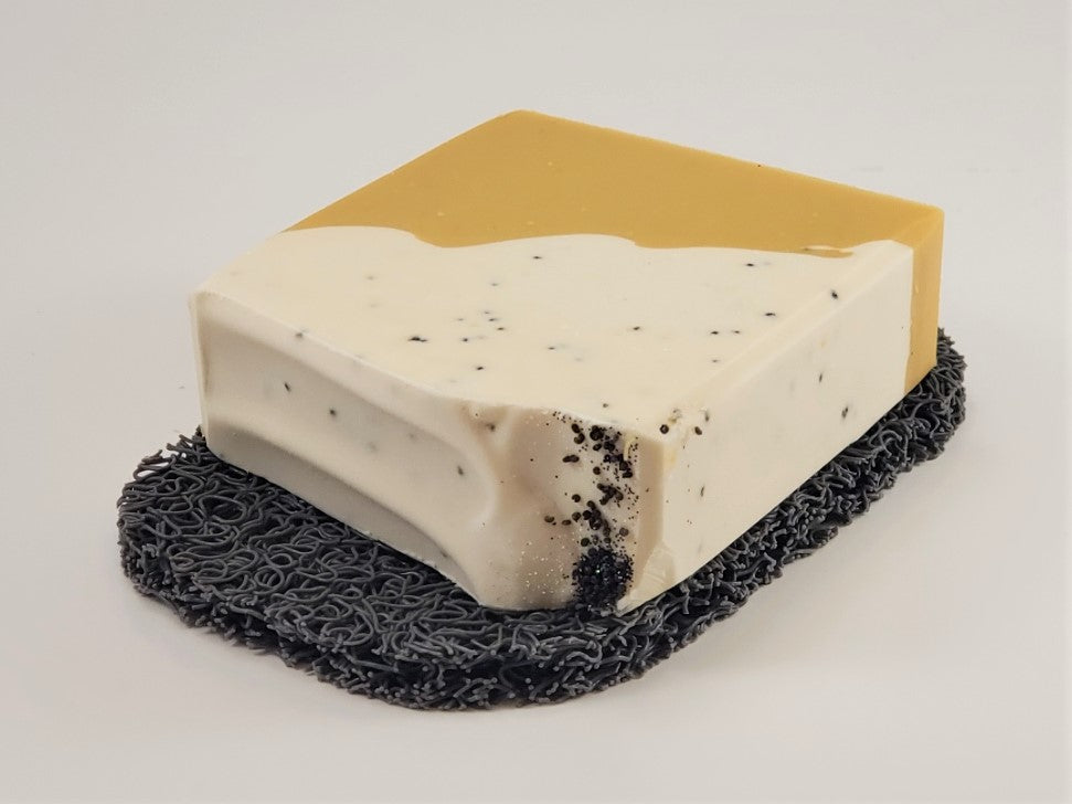 A handcrafted soap bar featuring a smooth beige top and a speckled white bottom rests elegantly on the Amillia Acres Soap Saver. This textured black dish is designed to extend the life of your soap, with its delicate particles enhancing both its functionality and aesthetic appeal.