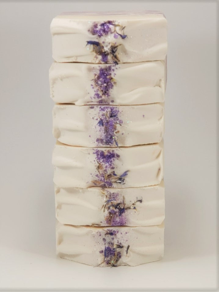 Grace by Amillia Acres is a handmade goat milk soap, cream-colored with a central stripe of dried lavender flowers and herbs. Its smooth texture features slight ridges, accentuated by natural oils for an artisanal look.