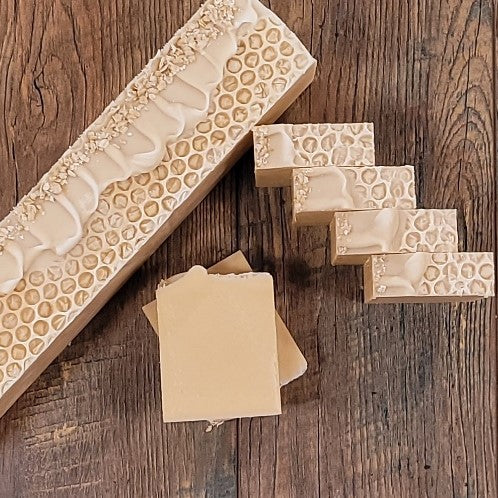Rectangular soap bars are displayed on a wooden surface. One large uncut bar, made with natural oils, features textured patterns, while five smaller handmade bars are aligned beside it. Two Nourish Goat Milk Soaps by Amillia Acres are laid flat in the foreground, revealing a smooth surface and light beige color.