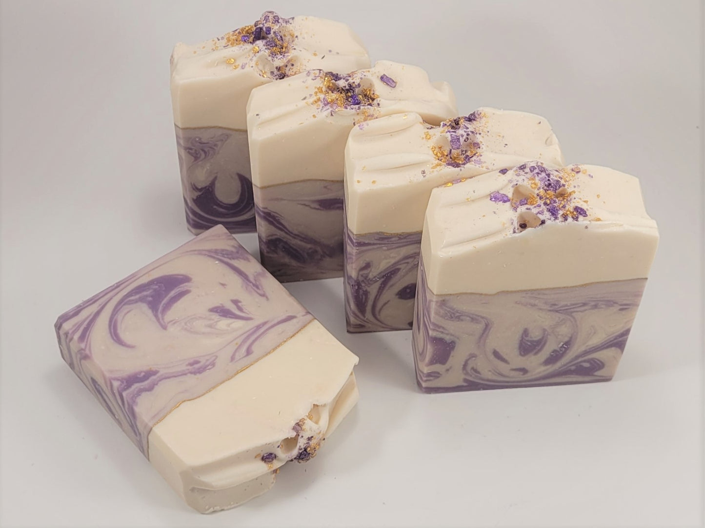 Five Grace goat milk soaps by Amillia Acres, in cream and purple swirls, are displayed. Four stand upright displaying a textured top with bits sprinkled on it, while one lies flat. These handmade lavender soaps have an appealing marbled pattern against a white background.