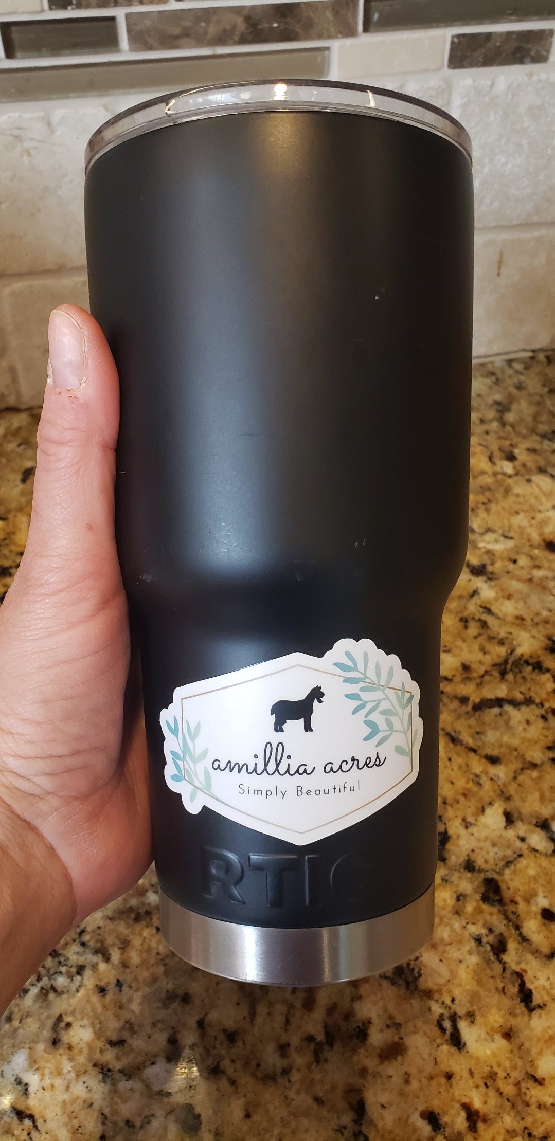 A hand holds a black insulated tumbler featuring an "Amillia Acres ~ Goat Milk Soaps and Lotions" vinyl sticker with a horse silhouette. The durable label contrasts against the speckled countertop, highlighted by the tiled backsplash.