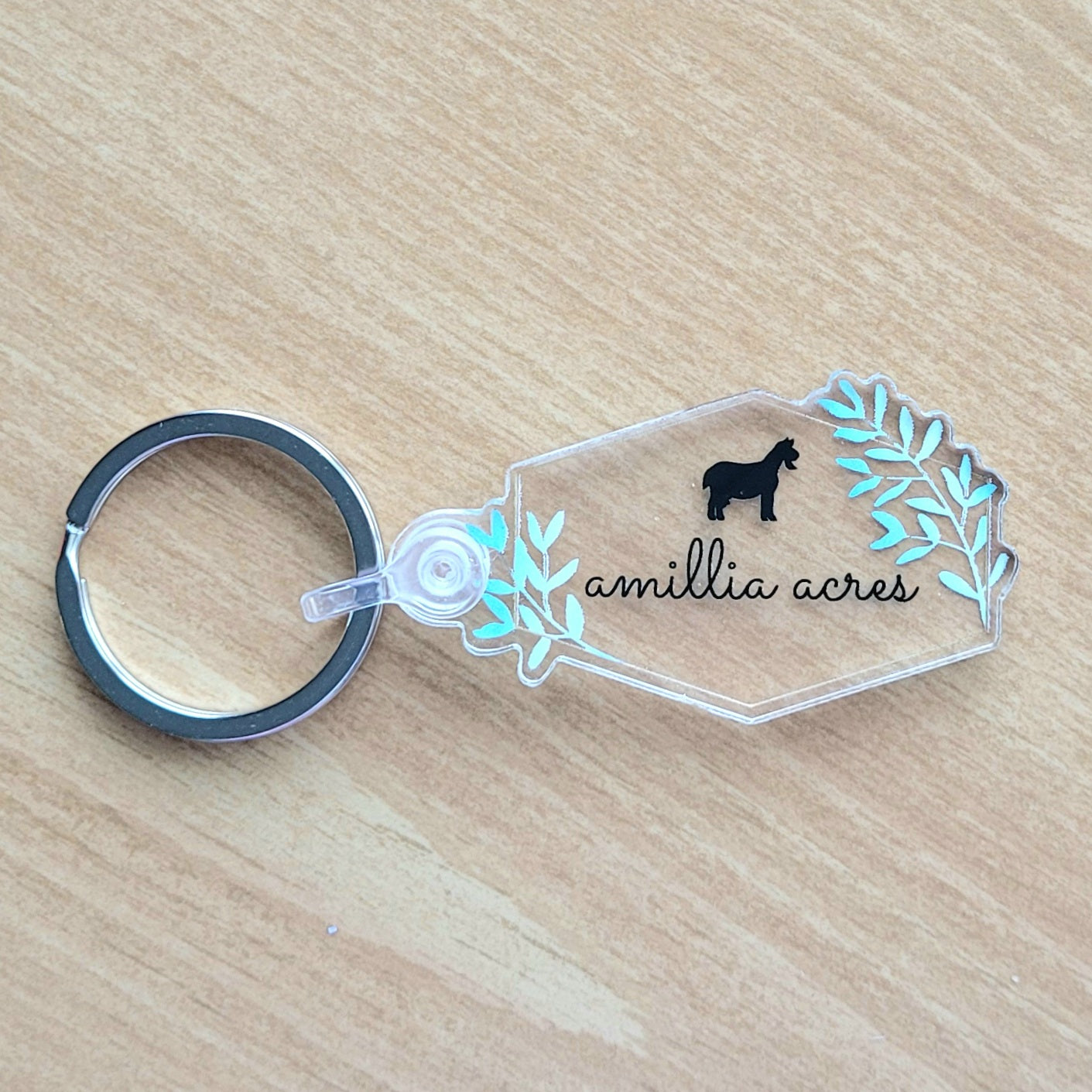 This product from Amillia Acres ~ Goat Milk Soaps and Lotions is a clear acrylic keychain featuring the name "Amillia Acres" in black script, adorned with a goat silhouette and green leaves. The design is elegantly complemented by a stainless steel key ring, making it stand out against a light wooden surface.