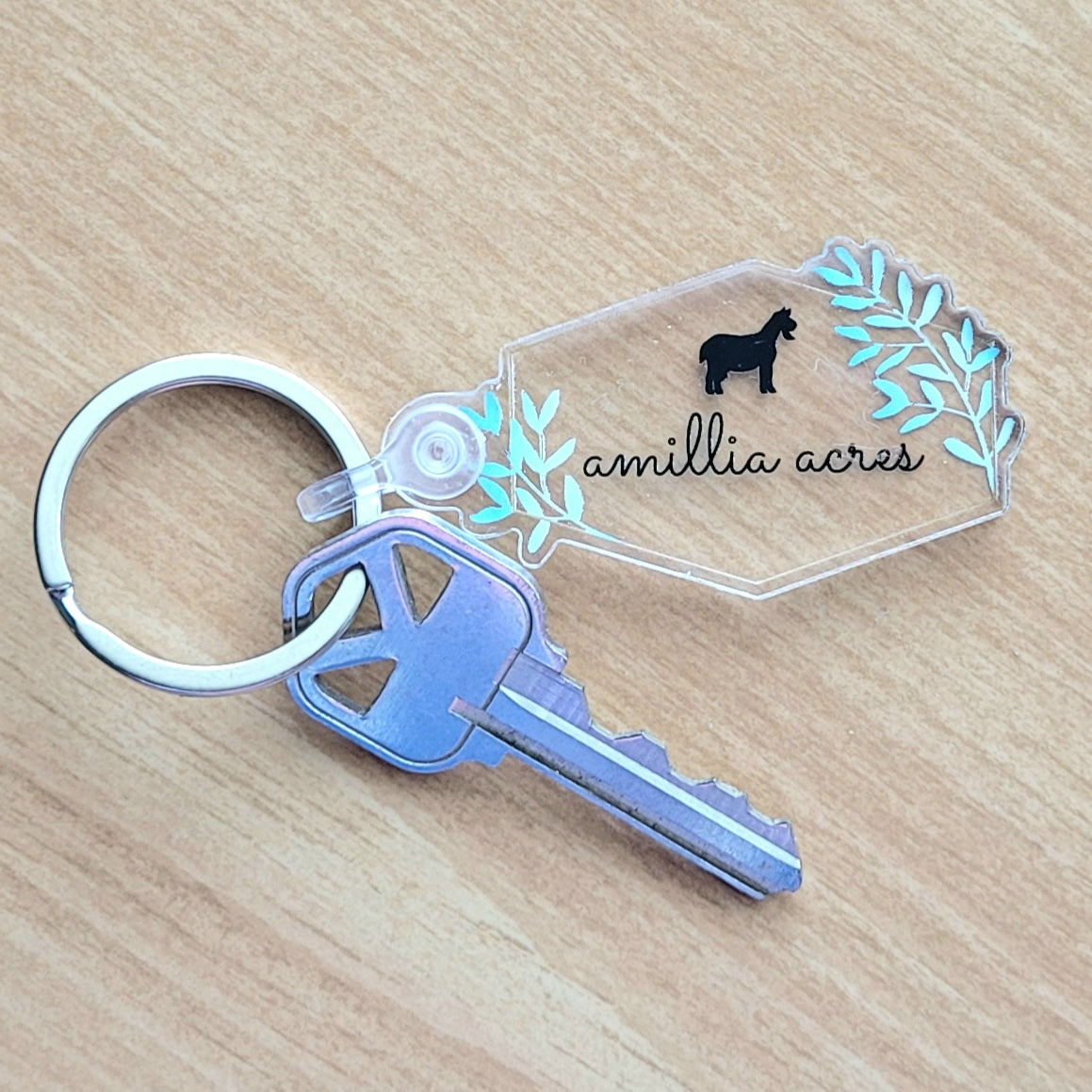 A refined key chain features a stainless steel ring securing a silver key, paired with a clear acrylic tag. The tag highlights a silhouette of a donkey with "Amillia Acres ~ Goat Milk Soaps and Lotions" engraved amidst leafy designs. These delightful pieces are gracefully displayed on a wooden surface.