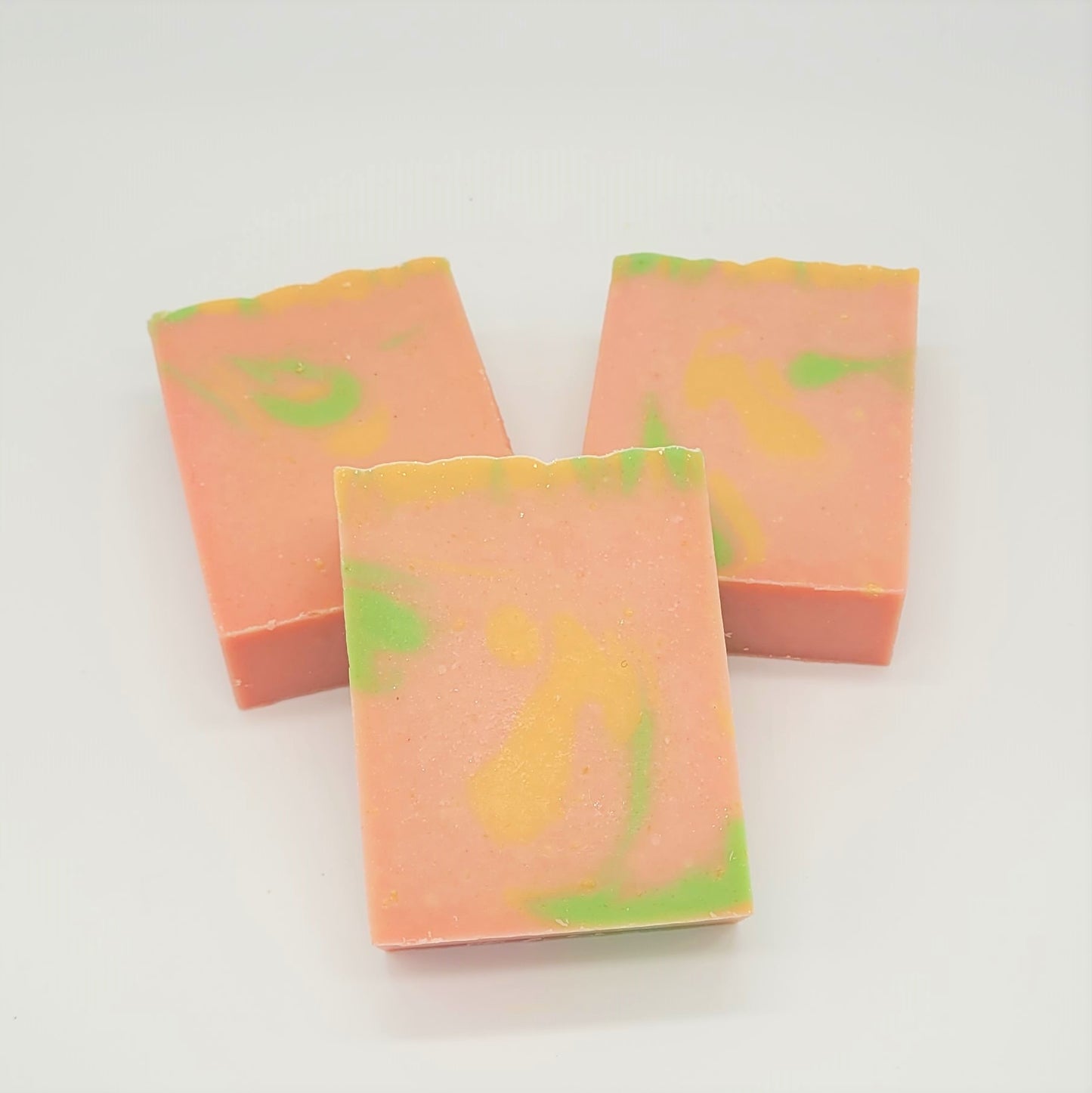 Three bars of Amillia Acres' "Happiness" goat milk soap, featuring a pink base with swirls of green and yellow, are arranged in a triangular formation on a plain white background.