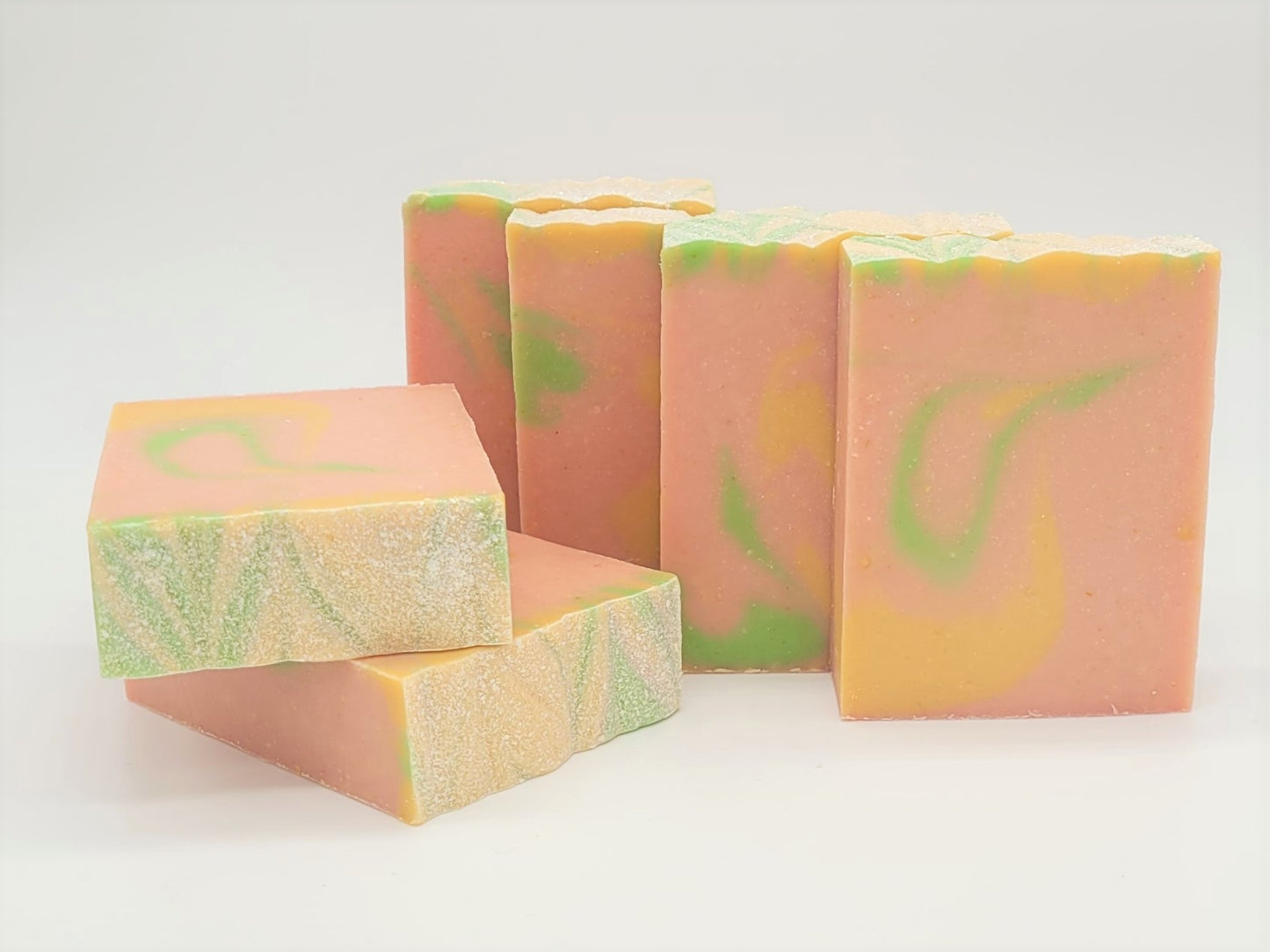 Five "Happiness" goat milk soaps by Amillia Acres feature green, yellow, and pink marbled swirls with textured tops. Made from natural oils, their shimmering surfaces stand out beautifully against a plain white background. These soaps are visually appealing and nourishing.