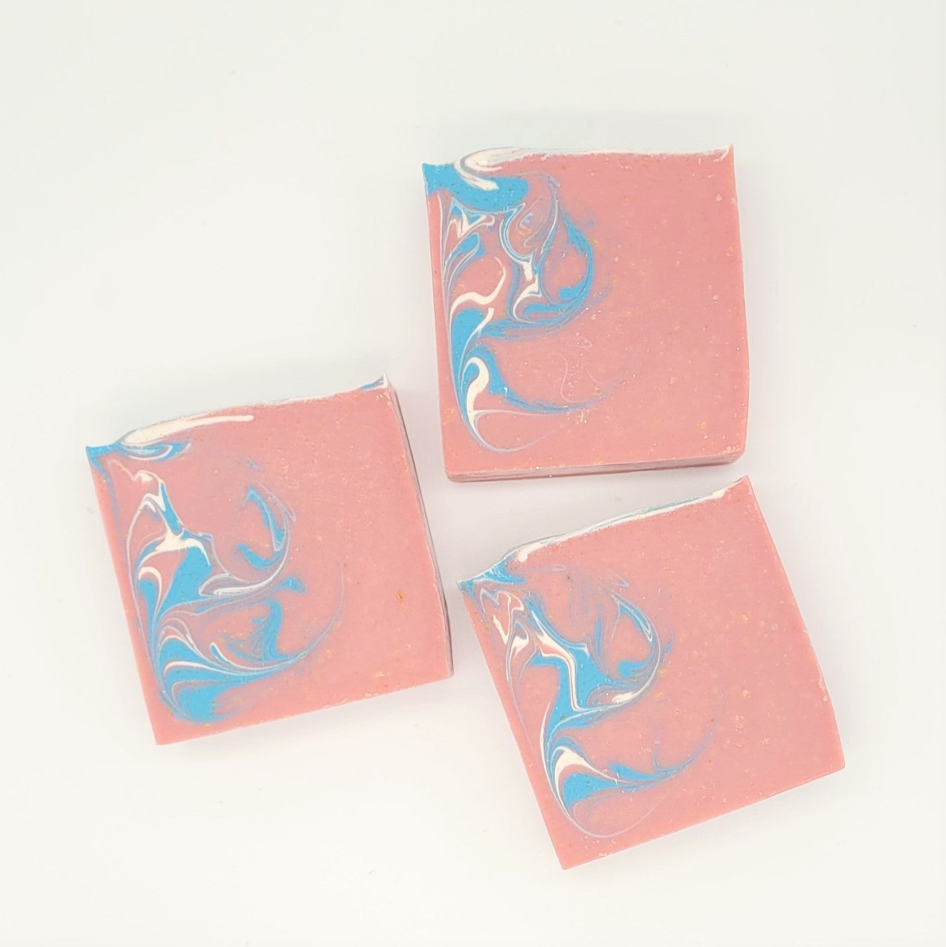 Three square pieces of Amillia Acres' Escape Goat Milk Soap, featuring swirled blue and white designs, are elegantly displayed on a white background. Their pink hue suggests a delicate blend of fragrances for a luxurious sensory experience.