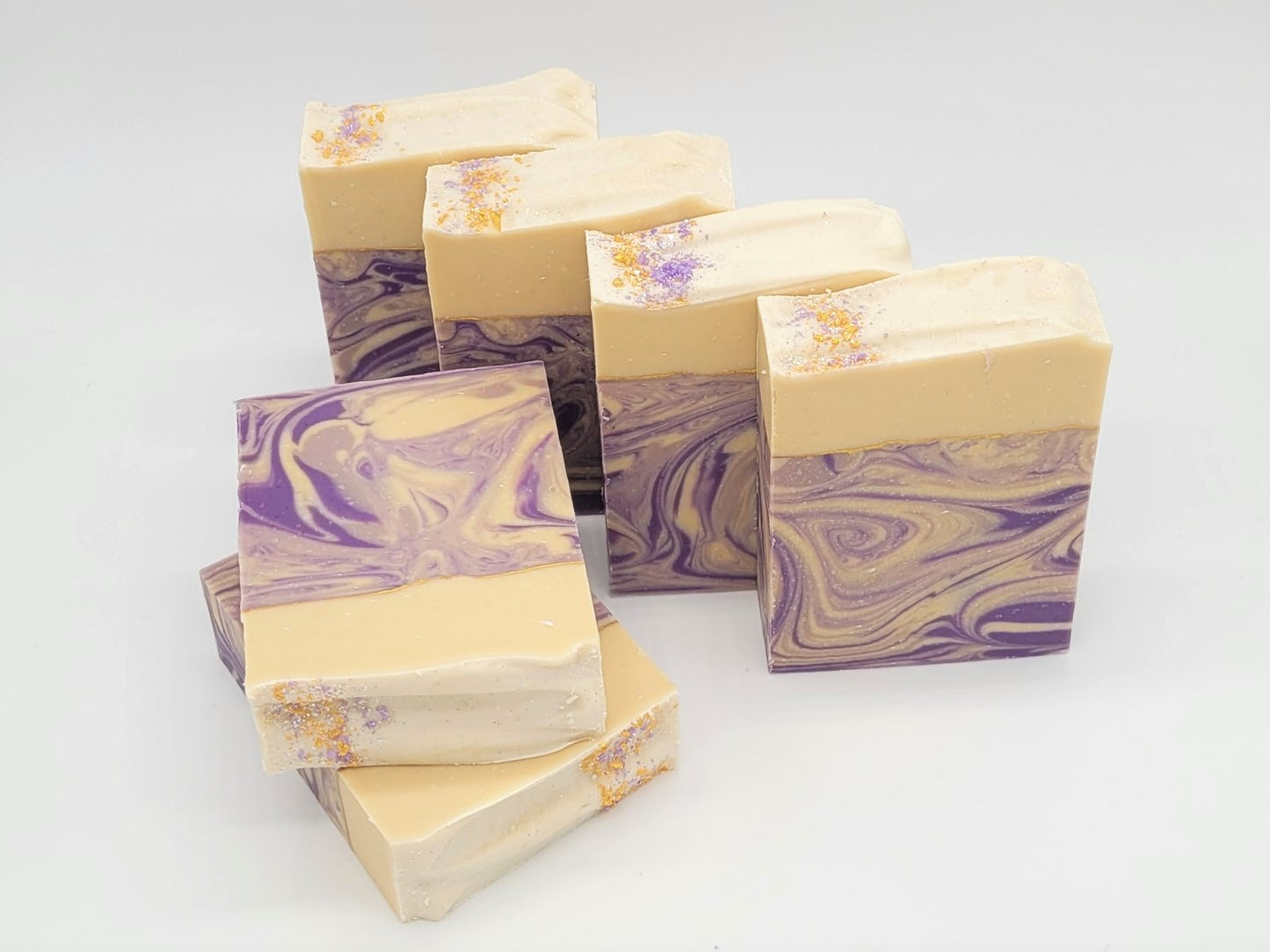 Six Grace goat milk soaps by Amillia Acres, featuring a cream and purple marbled design, are arranged in a staggered format on white. They are infused with natural oils, and their top edges have a dusting of purple and gold pigments, hinting at the lavender scent within.