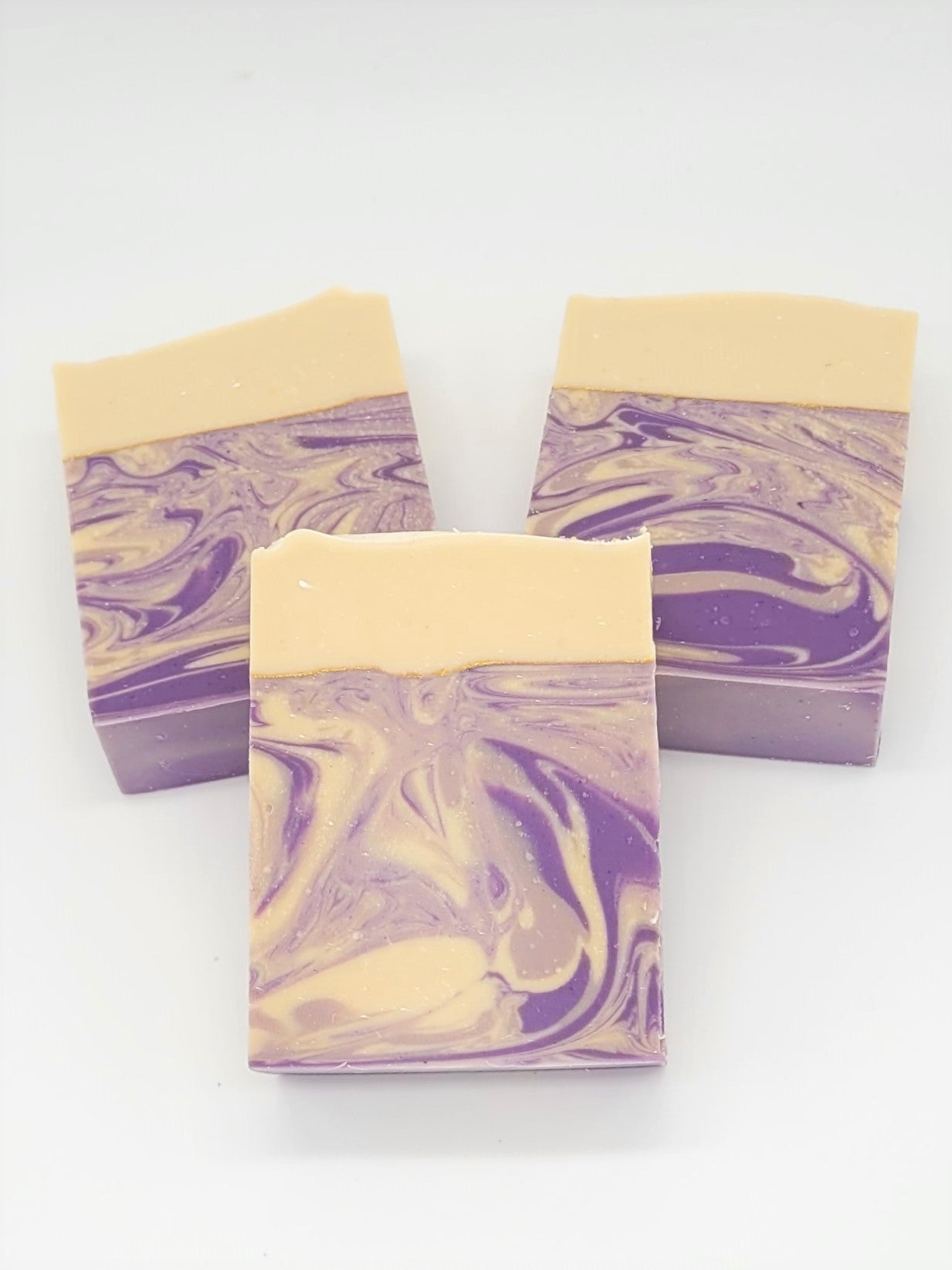 Three handmade rectangular soaps from Amillia Acres called "Grace" feature a swirled purple and cream design. Each lavender goat milk bar has a solid cream top, contrasting with the marbled pattern below, and is infused with natural oils resting elegantly on a plain white surface.