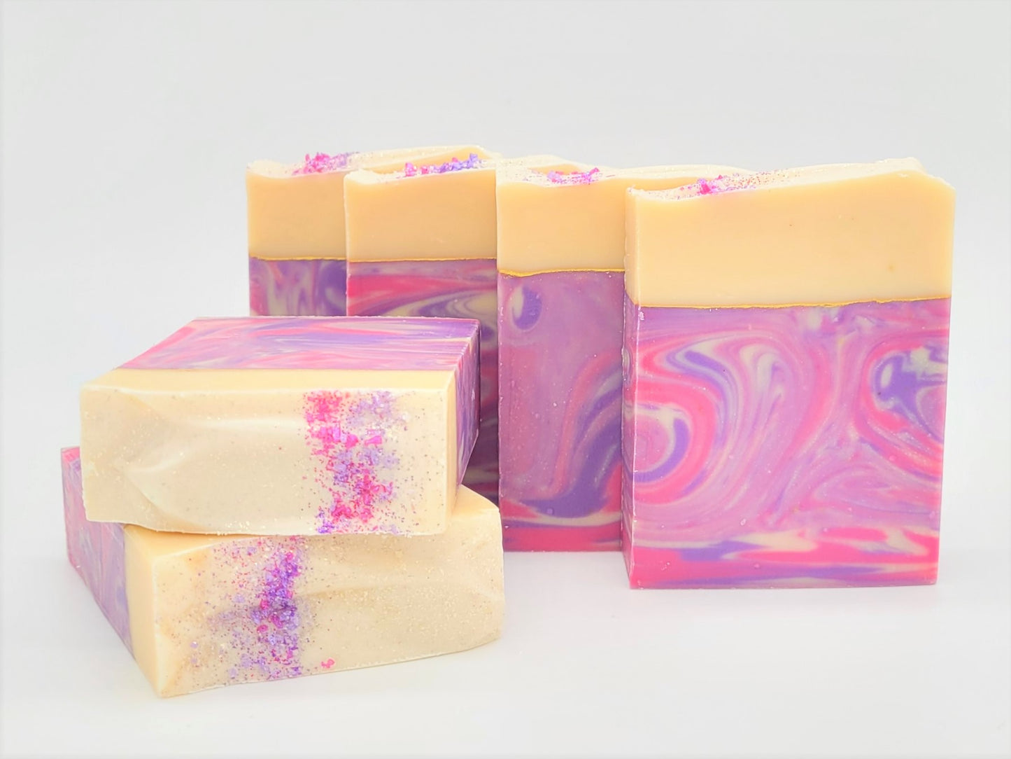 Several bars of Dazzle Goat Milk Soap by Amillia Acres, featuring marbled pink and purple swirls with a cream top, are arranged on a white background. These handmade soaps are infused with natural oils and adorned with pink and purple glitter accents.
