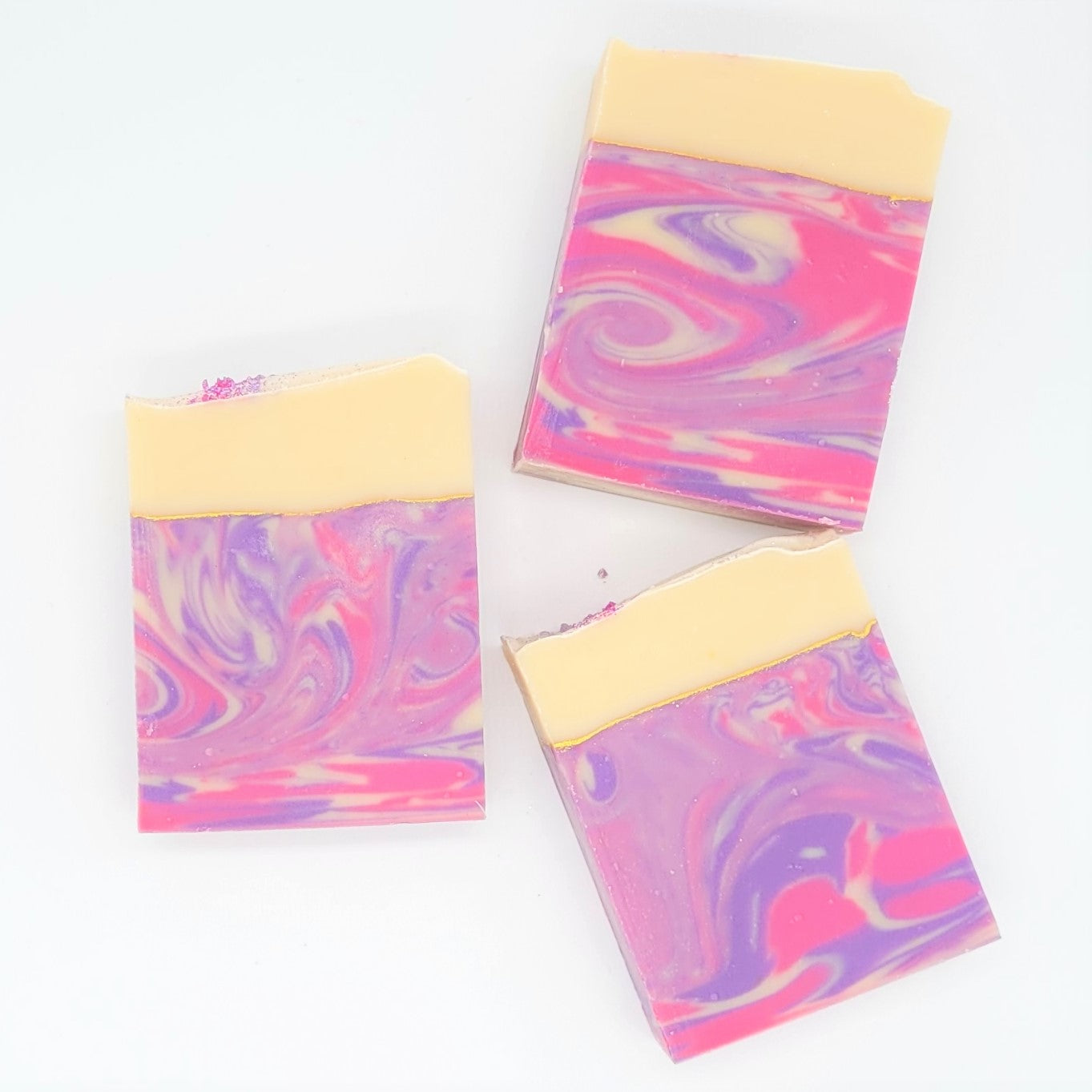 Three Dazzle Goat Milk Soap bars by Amillia Acres against a white background, each boasting a smooth, creamy top half and a marbled bottom with pink, purple, and white swirls infused with natural oils.