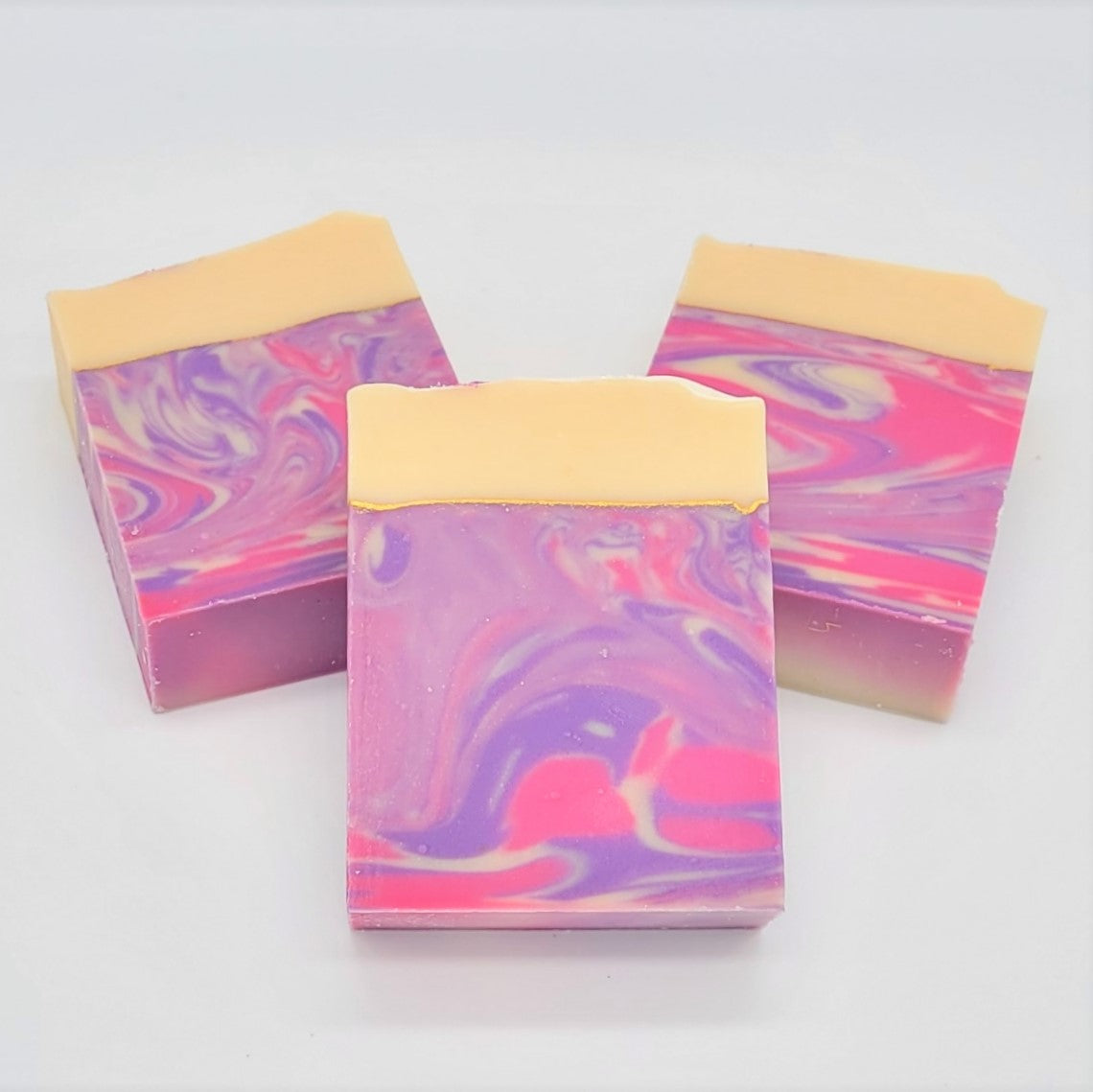 Three bars of Amillia Acres' Dazzle Goat Milk Soap, featuring a pink, purple, and white marbled design topped with a solid beige oil-enriched layer, are arranged on a plain white background.