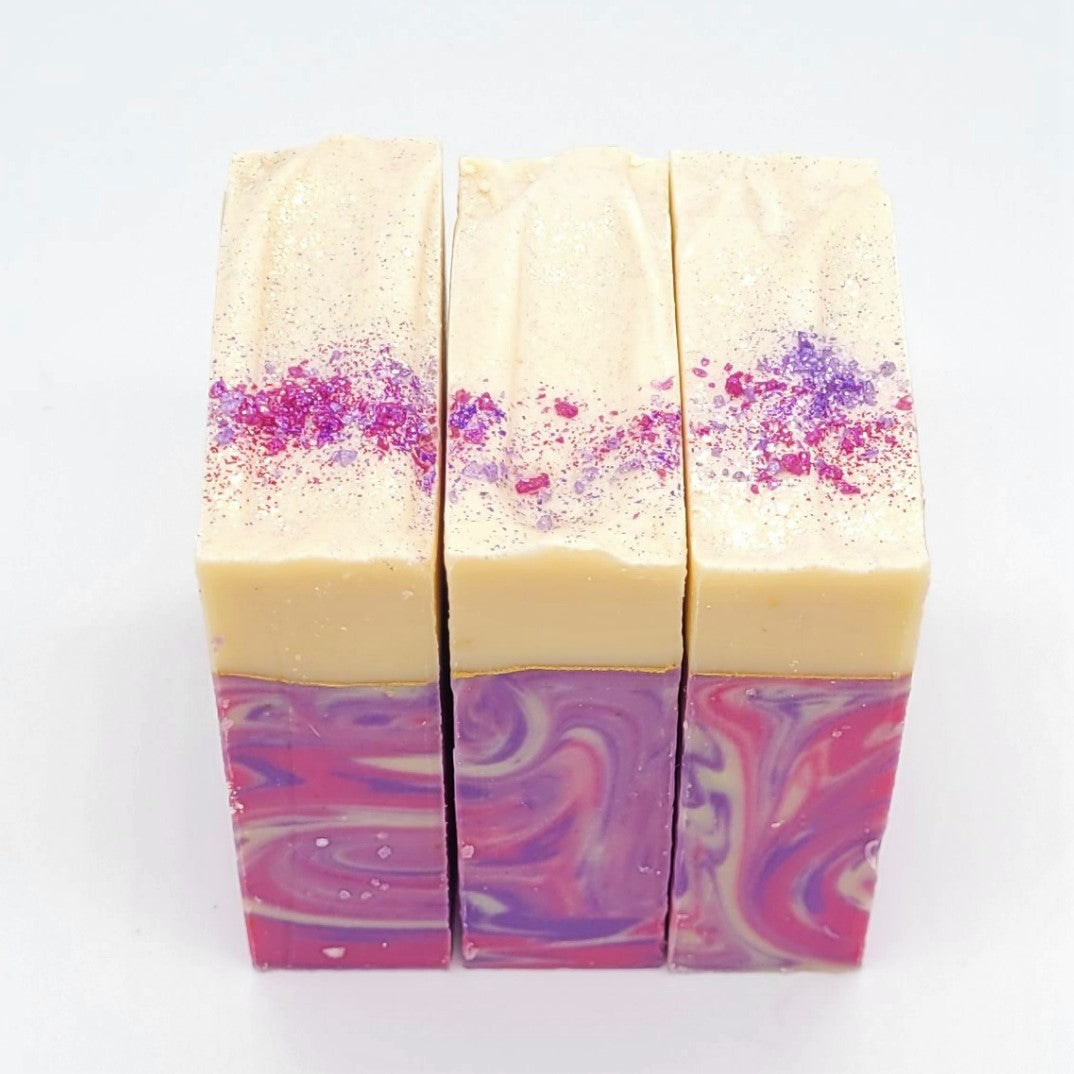 Three handcrafted Dazzle goat milk soaps by Amillia Acres feature creamy tops with pink and purple glitter, and the lower half showcases a swirling pink and purple pattern on a white background.