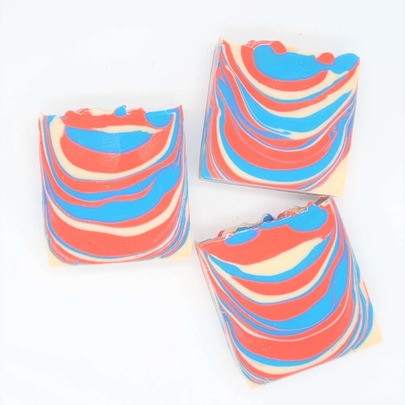 Three rectangular bars of Amillia Acres' Barbershop Goat Milk Soap, featuring vibrant red, blue, and cream swirls, are arranged on a white background. The marbled design resembles classic barbershop soap with an eye-catching and vivid effect.