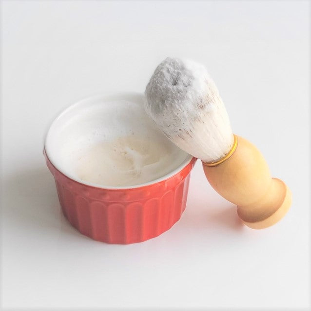 An Amillia Acres Shaving Soap Bar & Shave Set, featuring a red bowl filled with luxurious lather, rests on a white surface. Beside it, the wooden-handled brush, bristles rich in foam, leans gracefully against the bowl for the ultimate shaving experience with Goat Milk Soaps and Lotions.