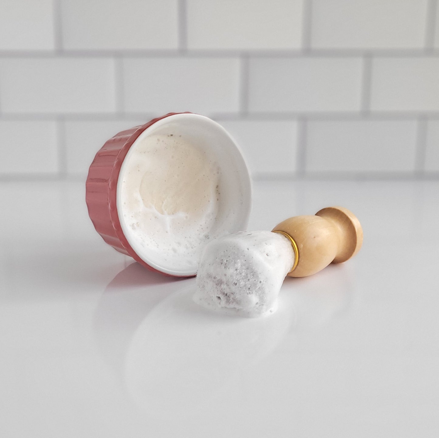 On a white surface, a luxurious shaving brush rests beside a vibrant red bowl filled with the rich lather of Amillia Acres ~ Goat Milk Soaps and Lotions' Shaving Soap Bar & Shave Set. The sleek white subway tiles in the background promise moisturized skin with each shave.
