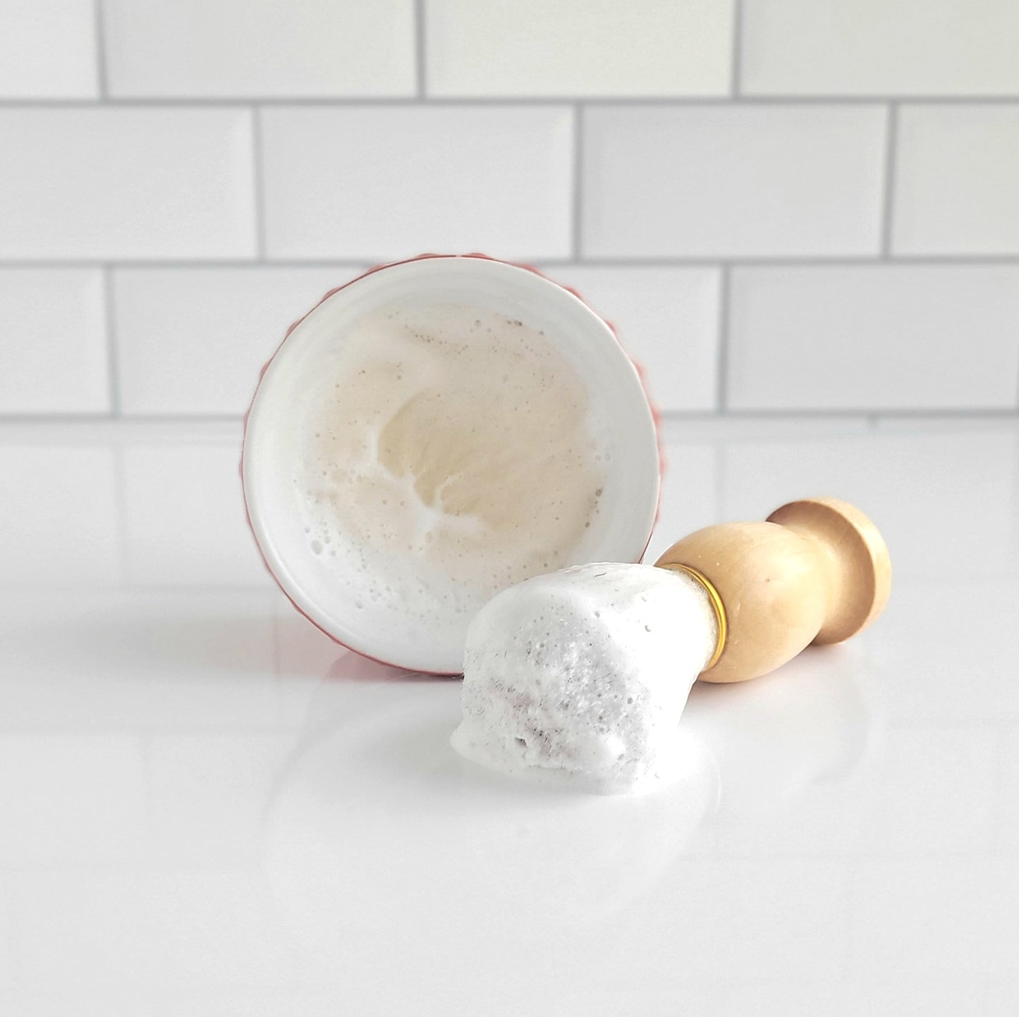 Nestled next to a bowl brimming with luxurious lather, the Shaving Soap Bar & Shave Set from Amillia Acres ~ Goat Milk Soaps and Lotions, featuring a shaving brush with a wooden handle, rests on a white tiled surface. This scene promises moisturized skin and an elevated shaving experience while exuding a clean, minimalist look.