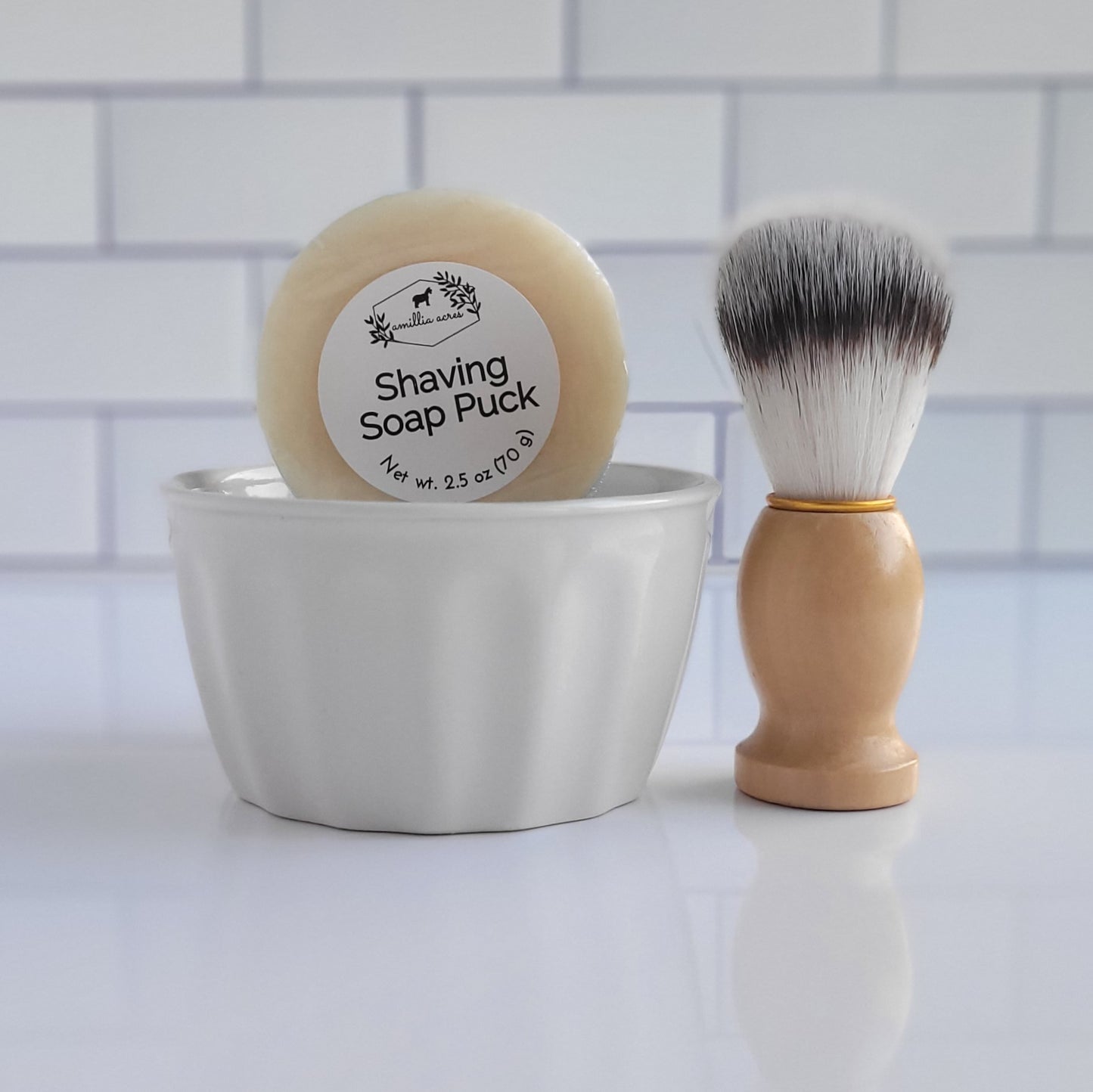 The Shaving Soap Bar & Shave Set by Amillia Acres ~ Goat Milk Soaps and Lotions includes a white ceramic bowl paired with a round shaving soap puck that guarantees a luxurious lather. Accompanying it is a shaving brush featuring a wooden handle and soft bristles, perfect for achieving moisturized skin. The background showcases classic white subway tile.