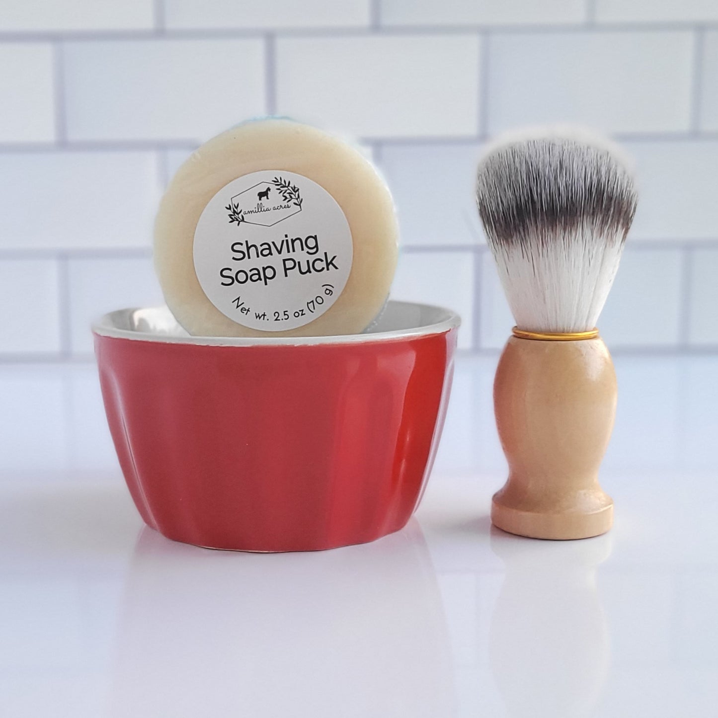 The "Shaving Soap Bar & Shave Set" by Amillia Acres ~ Goat Milk Soaps and Lotions includes a luxurious round soap bar in a red bowl, paired with a wooden-handled brush featuring white bristles. Its rich lather keeps skin moisturized, all beautifully showcased against a classic white subway tile backdrop.