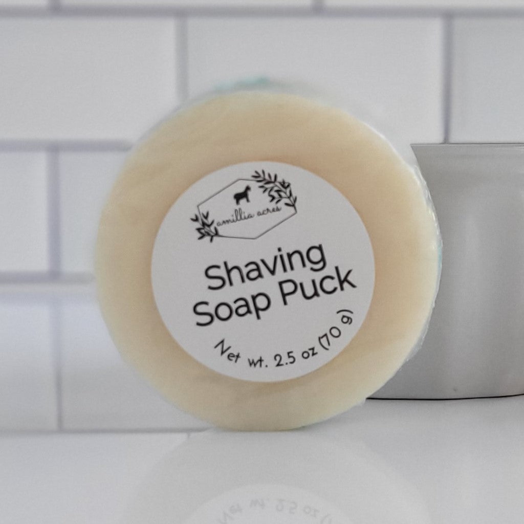 A close-up image of a shaving soap bar from the "Shaving Soap Bar & Shave Set" by Amillia Acres ~ Goat Milk Soaps and Lotions, with a label stating "Net wt. 2.5 oz (70 g)," promises a luxurious lather. The background features a white-tiled wall and a grey container, creating an ideal setting for an indulgent shave.