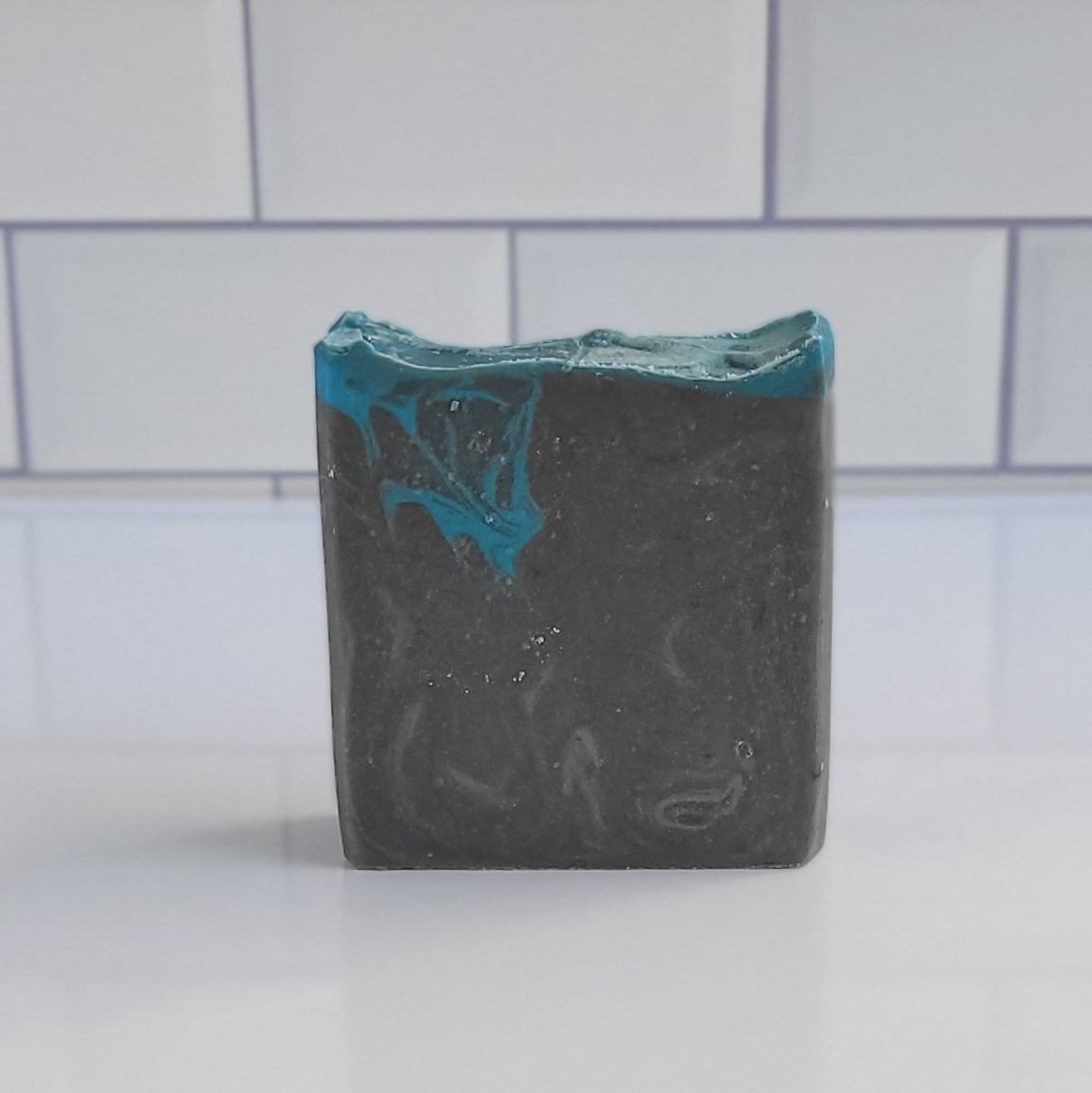 The Bay Rum Goat Milk Soap from Amillia Acres, with its marbled design in dark gray and teal, rests elegantly on a white countertop. Enriched with soothing goat milk, it complements the surrounding white subway tiles effortlessly.