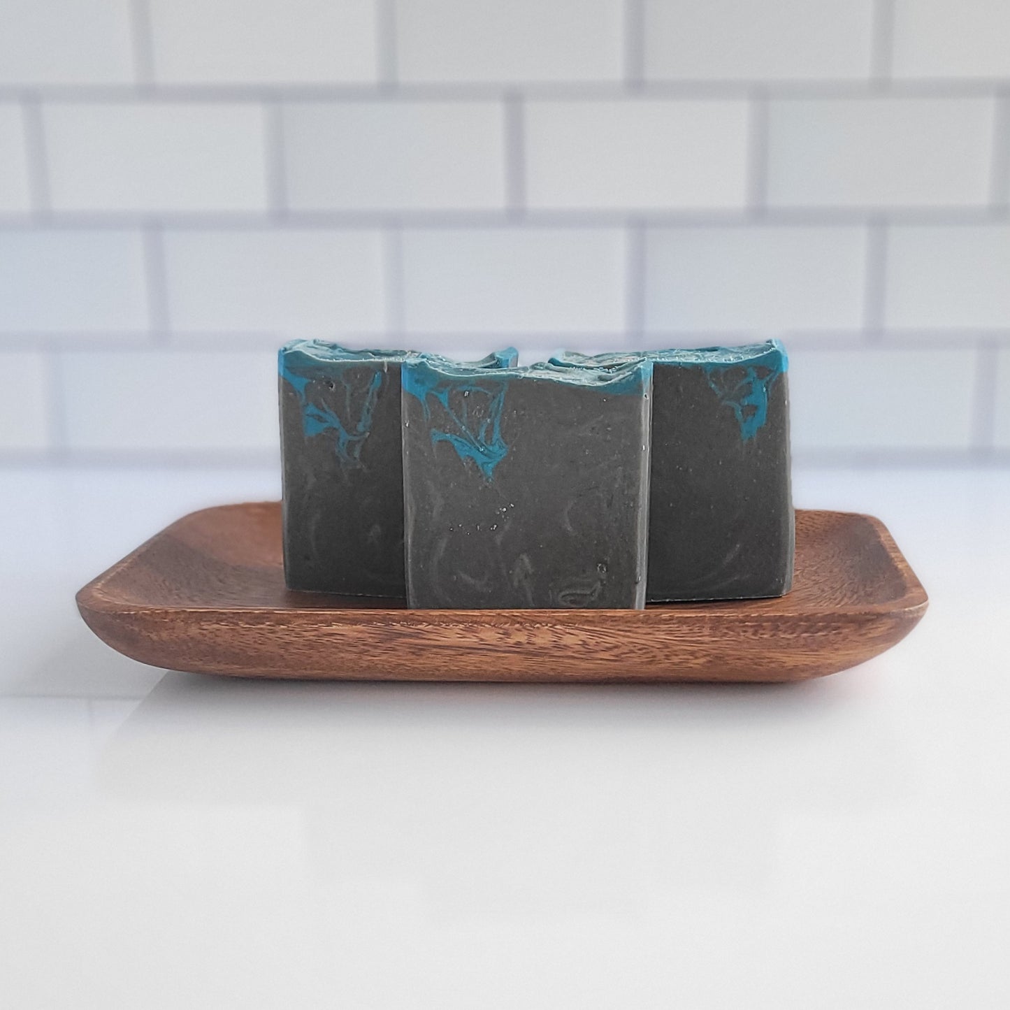 Three Bay Rum goat milk soap bars from Amillia Acres, featuring a dark base and turquoise swirls, are neatly placed on a wooden soap dish against a subway-patterned white tile backdrop.