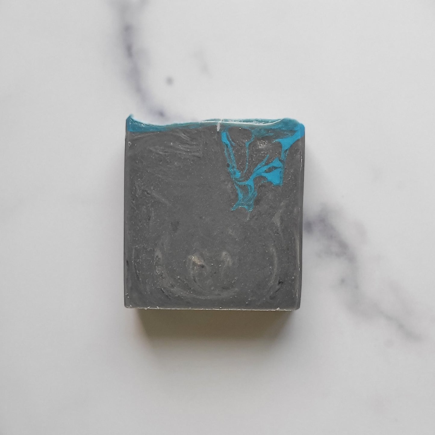 A square bar of Amillia Acres' Bay Rum Goat Milk Soap, featuring a marbled gray and turquoise design, rests on a white marble surface. Infused with goat milk, it showcases swirls of color and a smooth texture.
