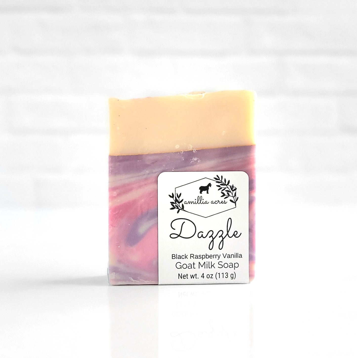 The Amillia Acres "Dazzle" goat milk soap, scented with black raspberry vanilla, weighs 4 oz (113 g) and is enriched with natural oils. It's handmade with a swirl of pastel purple, pink, and cream colors on a white backdrop.