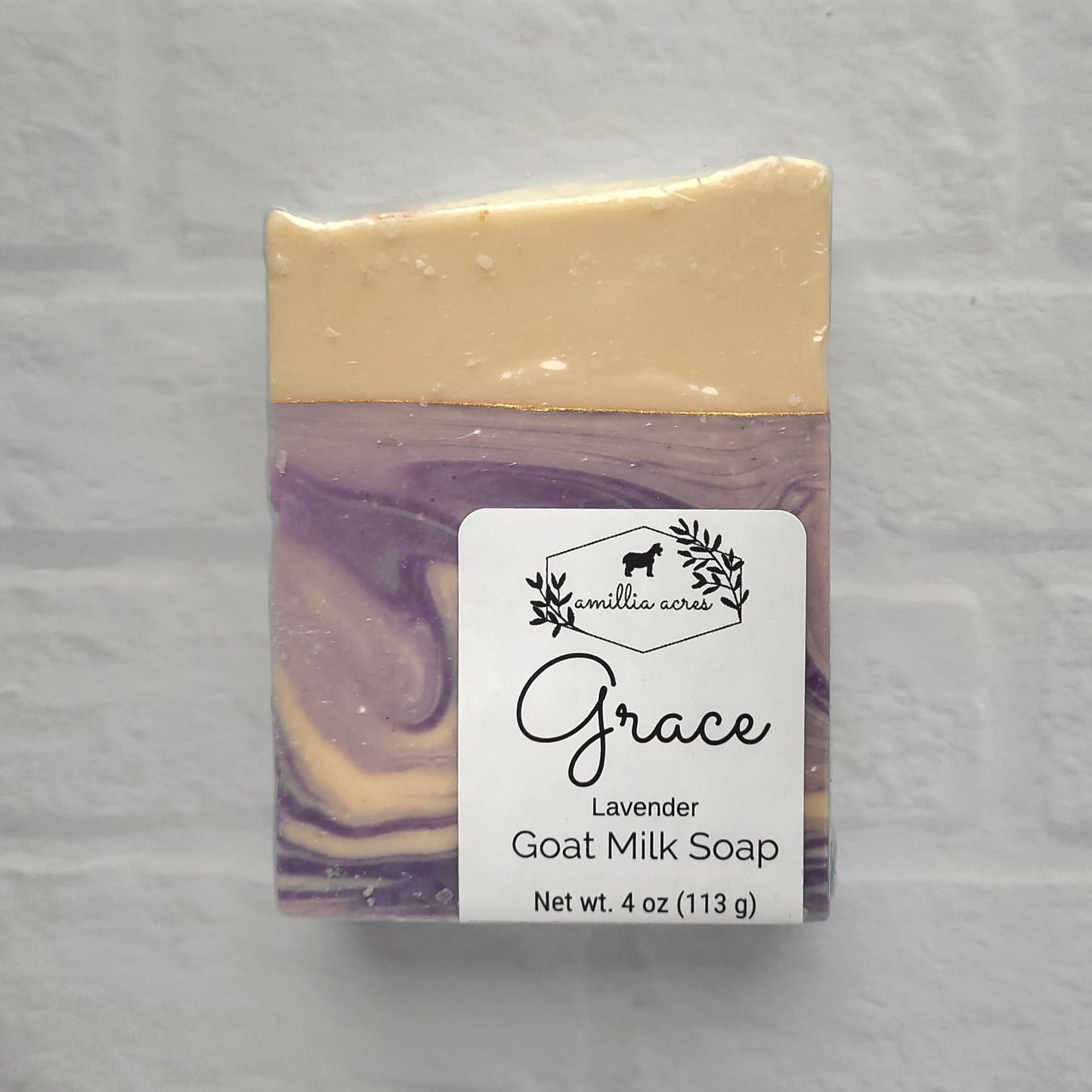 A square bar of Amillia Acres' Grace Goat Milk Soap, featuring purple and cream swirls, has a label with a goat and leaves stating "Grace Lavender Goat Milk Soap, Net wt. 4 oz (113 g)." Infused with nourishing natural oils, it is displayed against a white brick wall backdrop.