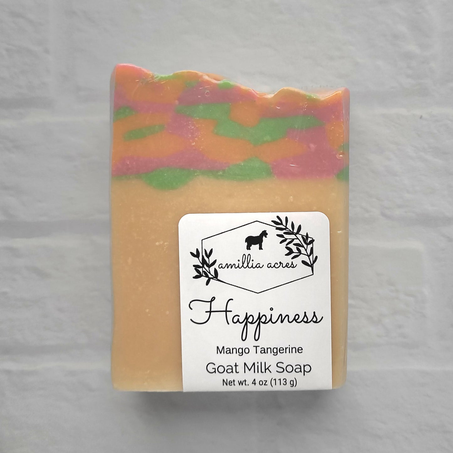 A bar of "Happiness" goat milk soap from Amillia Acres features a mango tangerine scent, colorful layers of pink, orange, and green, and is handmade with natural oils. The label depicts leaves and a horse silhouette, enhancing its artisanal charm.