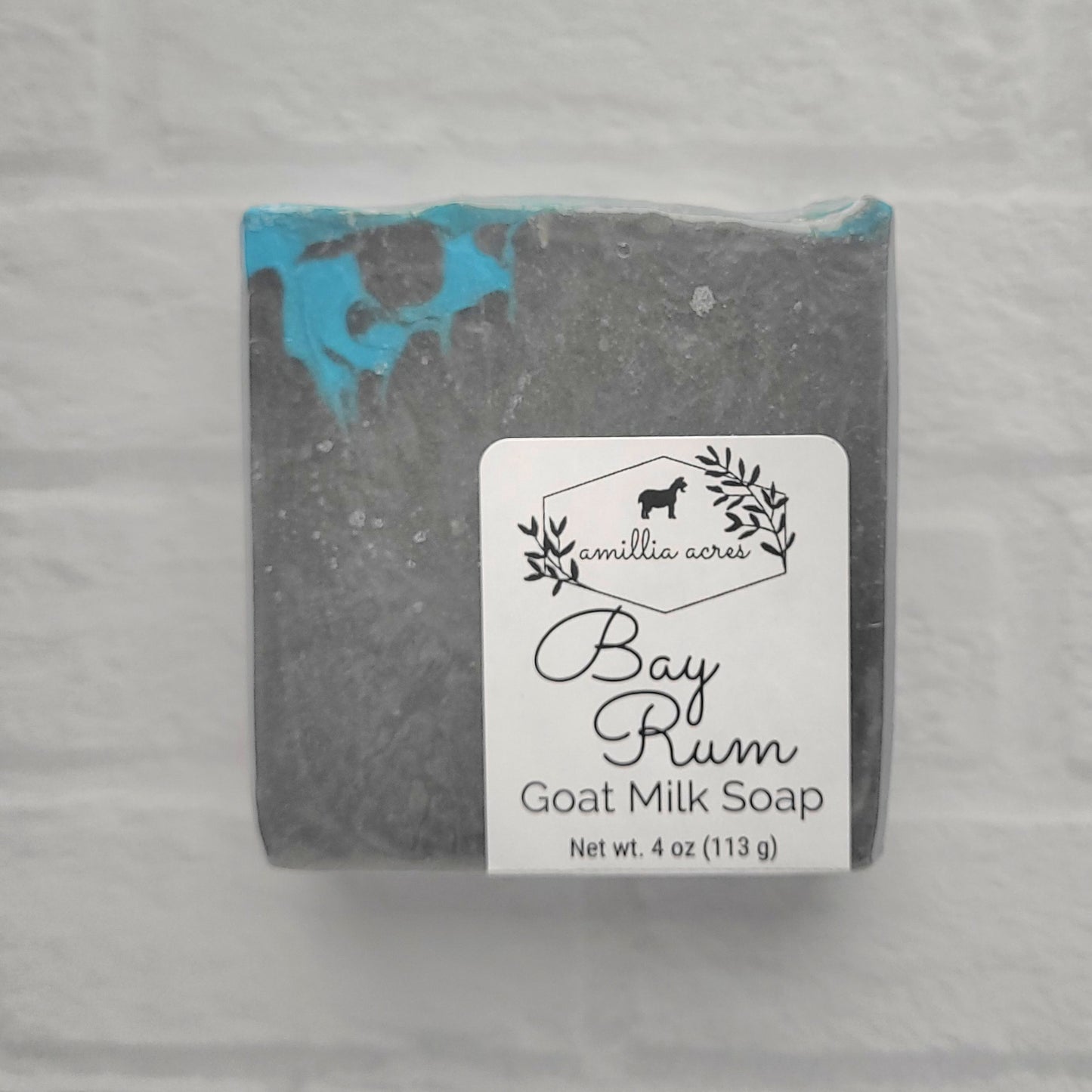 The Amillia Acres Bay Rum Goat Milk Soap, weighing 4 oz (113 g), features a marbled blue and gray design. Its label, adorned with a goat and leaves, showcases the careful craftsmanship as it rests on a textured white background.