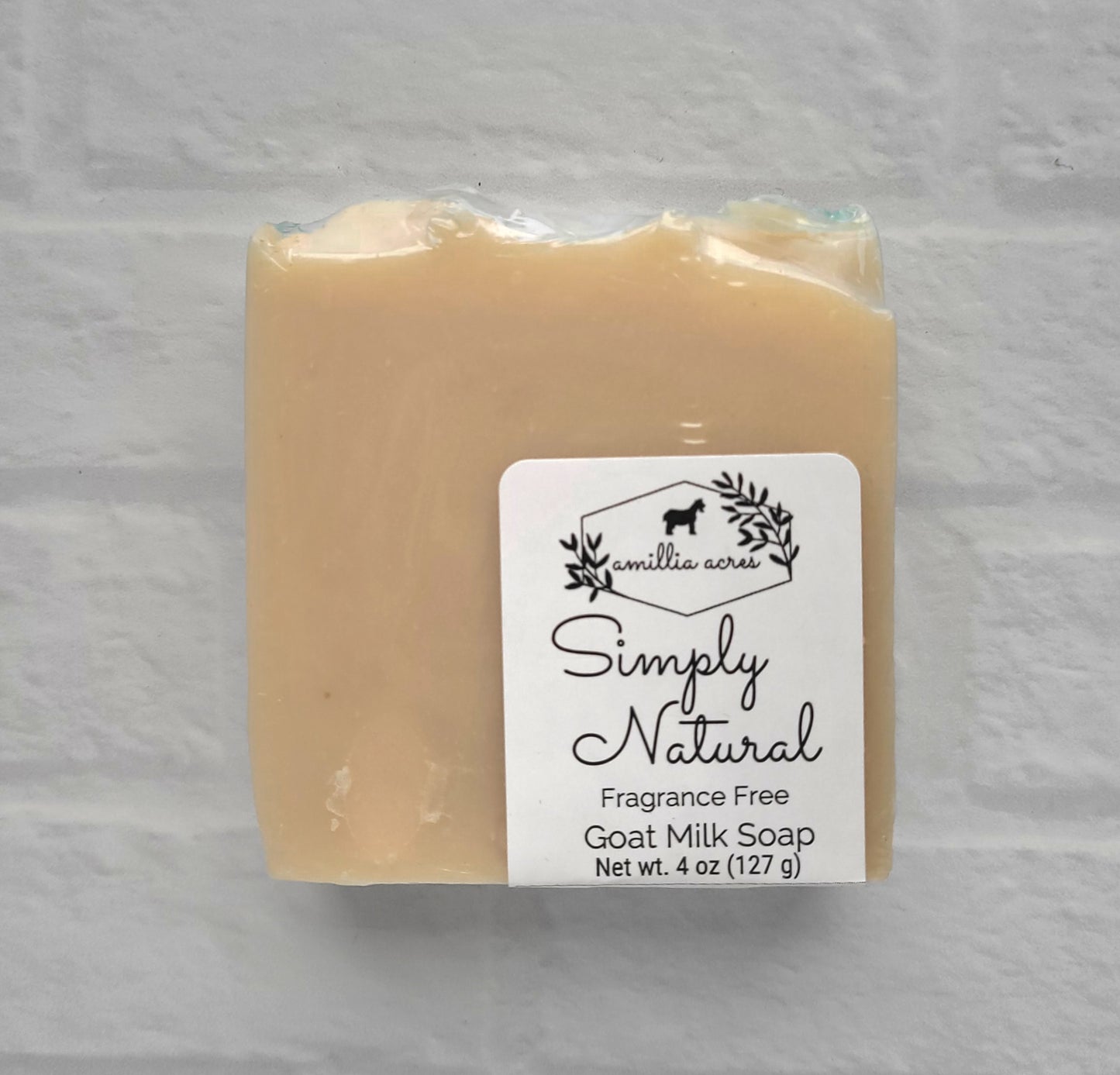 A beige, fragrance-free handmade goat milk soap bar, labeled "Amillia Acres Simplicity," weighing 4 oz (127 g), rests against a light gray brick background.