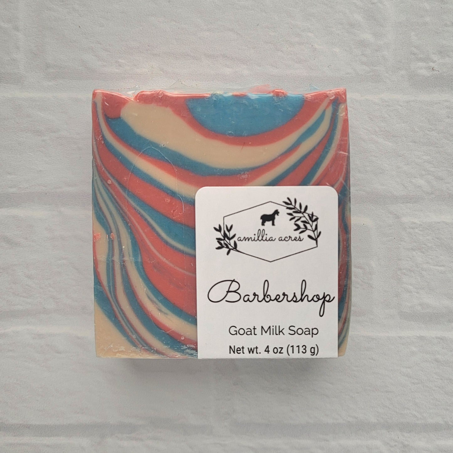 A handmade goat milk soap bar features swirling red, blue, and cream colors on a light, textured background. The label reads "Amillia Acres ~ Goat Milk Soaps and Lotions, Barbershop Soap, Net wt. 4 oz (113 g).